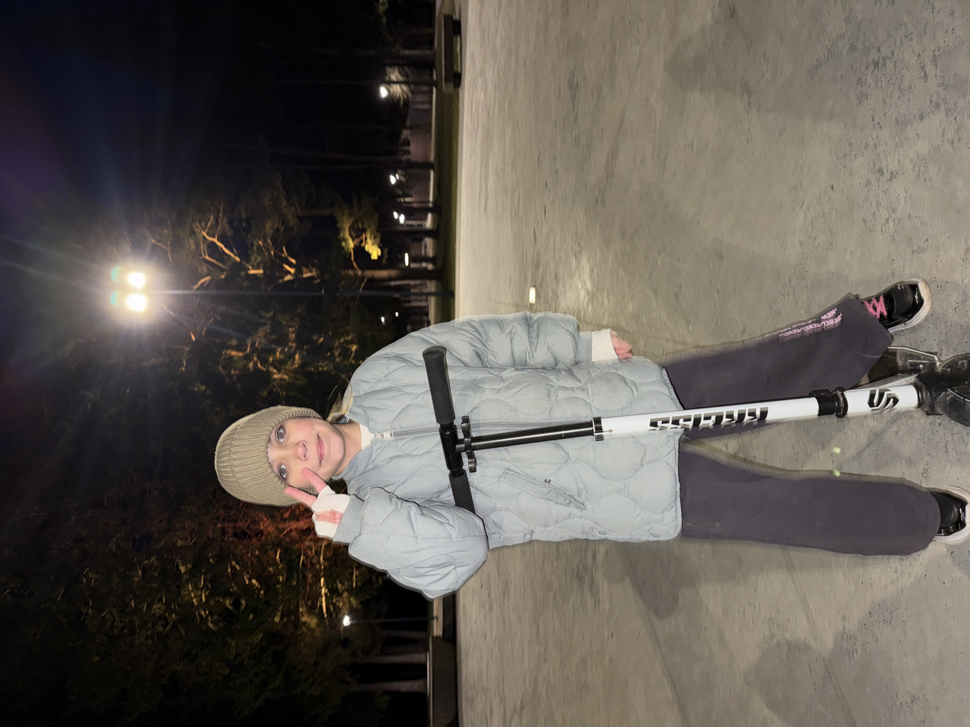 A young individual is captured standing upright at night on a flat, grey concrete surface, possibly a skate park or open plaza. They are wearing a light blue/grey quilted puffer jacket, dark grey pants with "REBEL REBEL REBEL" text faintly visible along the side, black sneakers with bright pink laces, a light-colored knit beanie, and white fingerless gloves or mittens. The person is holding a white and black stunt scooter, with "KRISS" printed vertically on its handlebar stem. They are facing slightly towards the camera, smiling, and making a peace sign with their left hand.
The background is dark, indicating it is nighttime. Several bright streetlights illuminate the scene, casting light on some distant trees, which appear silhouetted. The overall atmosphere suggests a cool or cold evening, given the winter-appropriate attire of the person. The lighting creates bright flares from the streetlights. The person appears happy and engaged, likely enjoying an evening outing with their scooter.