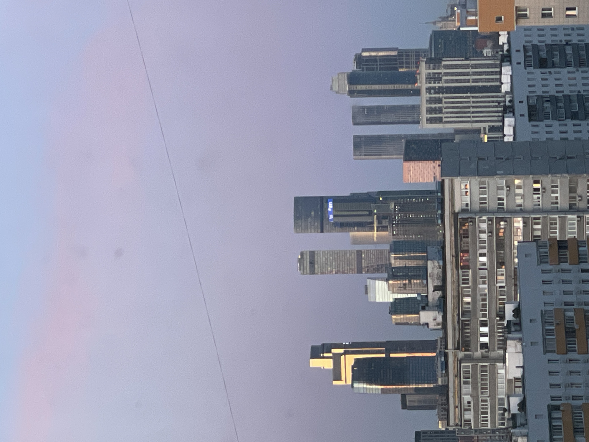 The image captures a panoramic view of a city skyline in Khimki, Russia, at twilight, either during a late sunset or early sunrise. The sky, which occupies the left two-thirds of the frame, displays a soft gradient of purple, pink, and light blue hues, indicating the transitional lighting conditions. A thin, dark line, likely a power line or cable, stretches horizontally across the upper left portion of the sky.
The right side of the image features a dense cluster of urban buildings, showcasing a mix of architectural styles. Tall, modern skyscrapers with reflective glass facades stand alongside more traditional multi-story residential or office buildings, characterized by numerous rectangular windows and balconies. Many of these windows are illuminated, casting a warm glow across the scene and suggesting active internal spaces. Some of the distant skyscrapers also display indistinct blue glowing signs or corporate logos on their upper levels, though no specific text is clearly legible.
No people or specific activities are discernible within the frame, but the illuminated buildings and dense urban landscape imply a vibrant city. The weather appears clear and calm, with no clouds visible. The scene portrays a tranquil yet expansive urban environment during the quiet hours of twilight.