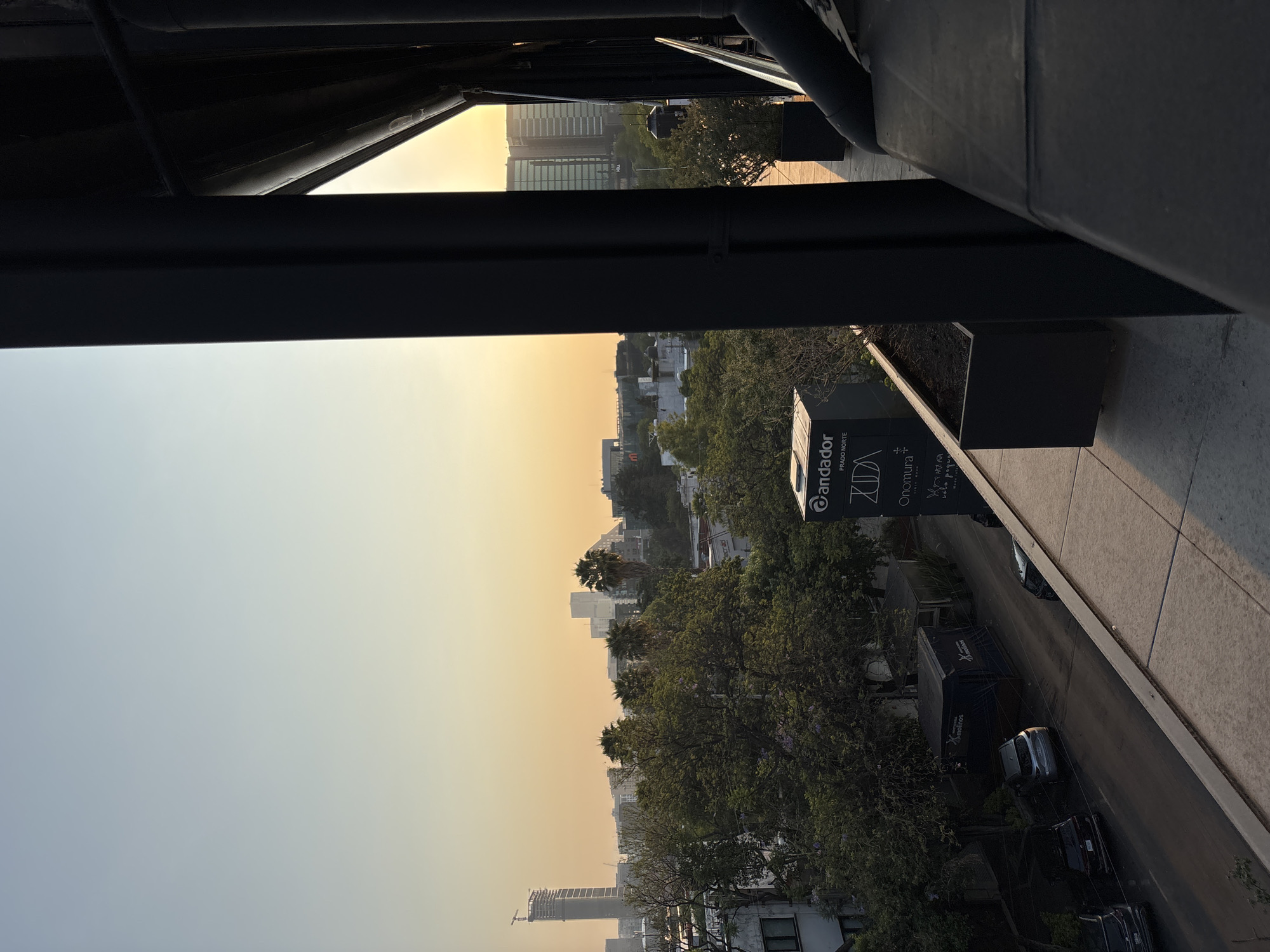 The photograph captures a cityscape, likely taken from an elevated position such as a balcony or high-rise building, overlooking an urban landscape in Mexico City, Mexico.
The scene is characterized by a mix of residential and commercial buildings, interspersed with greenery from trees. The sky is a gradient of colors, hinting at either sunrise or sunset.
In the foreground, architectural elements of the building from which the photo was taken, including a dark metal structure.
A street is visible with parked and moving vehicles. A sign for "andador prado norte" is also visible.