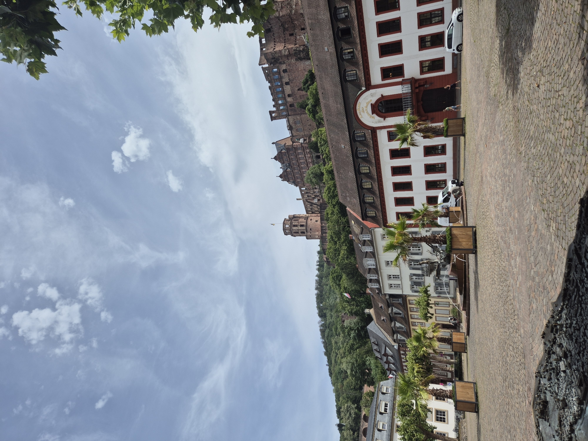 This is a vertical, eye-level, outdoor shot of a European town square with a castle visible on a hill in the background. In the foreground, there is a cobblestone square with planters holding palm trees and a fountain. Behind the planters are buildings with white facades, red trim, and dark windows. To the left, a white van is parked. To the right, a white car is parked. Two people are standing in front of the building on the right, and one person is walking a dog. In the middle ground, a row of older buildings with gray roofs runs parallel to the square. In the background, a large castle complex with stone towers and walls sits atop a lush, tree-covered hill under a partly cloudy blue sky. Green foliage from trees frames the top right corner of the image.