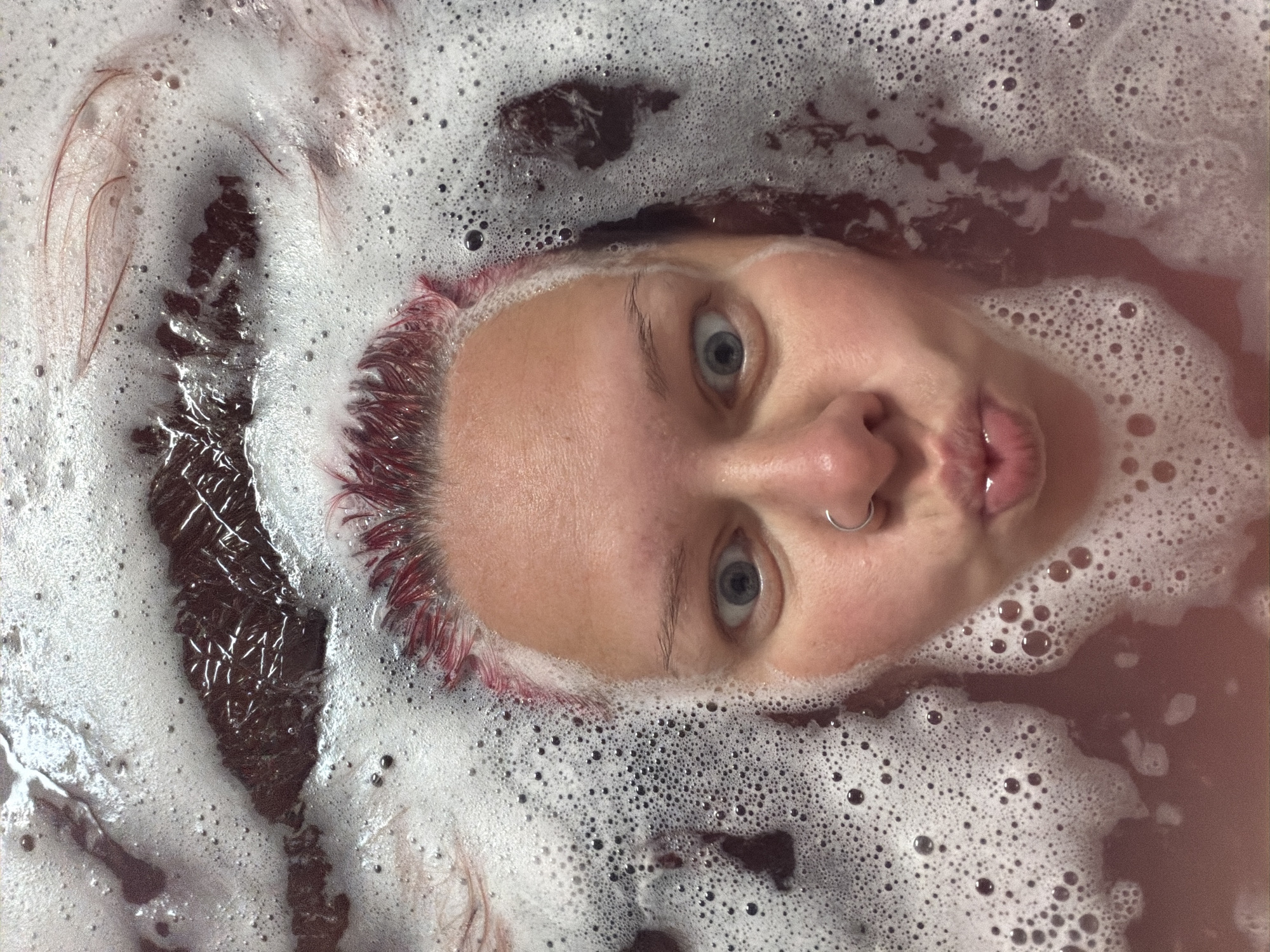 A close-up, overhead shot captures a person's face and upper body submerged in a bathtub filled with reddish-brown water and white foam. The person's eyes are wide open, and their lips are pursed, suggesting they are blowing a kiss or making a "fish face." They have bright pinkish-red hair, which is visible around their forehead and extending into the bubbly water. A silver hoop nose ring is visible on their right nostril. The foam obscures much of their body and the bathtub's contents, creating a textured, abstract background. The lighting is bright, illuminating the foam and the person's skin. The scene is suggestive of a relaxing or playful bath. There is no information in the image to indicate a specific location in Chaikovsky, Russia.