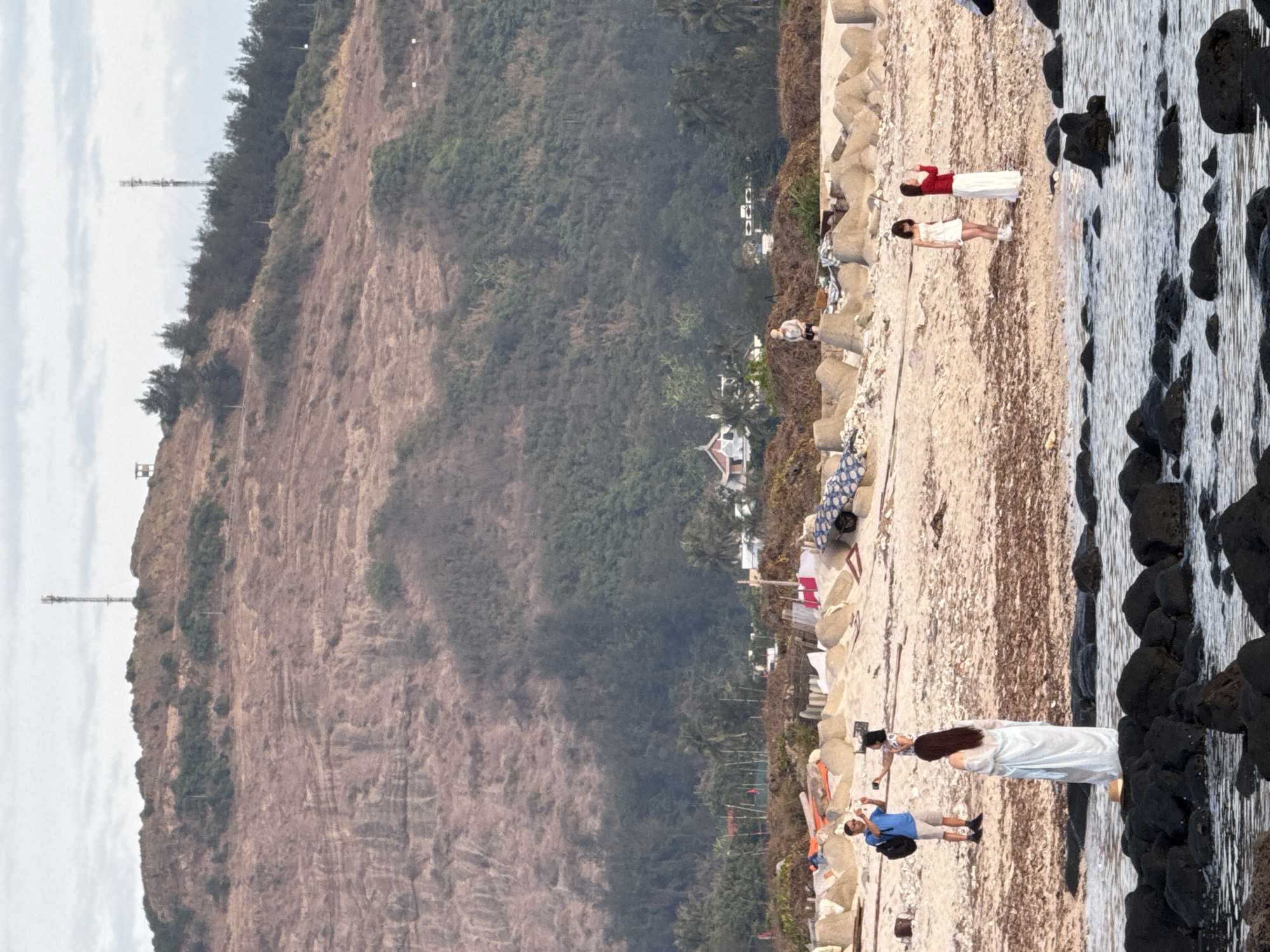 The image shows a beach on Lý Sơn Island, Vietnam. In the foreground, dark rocks are partially submerged in shallow water. A woman in a white dress stands on the rocks. Further back on the sandy beach, a man in a blue shirt and shorts is taking a photo of a woman standing next to him. In the middle ground, two women in white dresses are standing on the beach. Behind them, there are some debris, concrete barriers, and a thatched-roof structure. In the background, a green, tree-covered hill rises steeply, with a tall antenna tower visible at its peak. The sky is overcast.