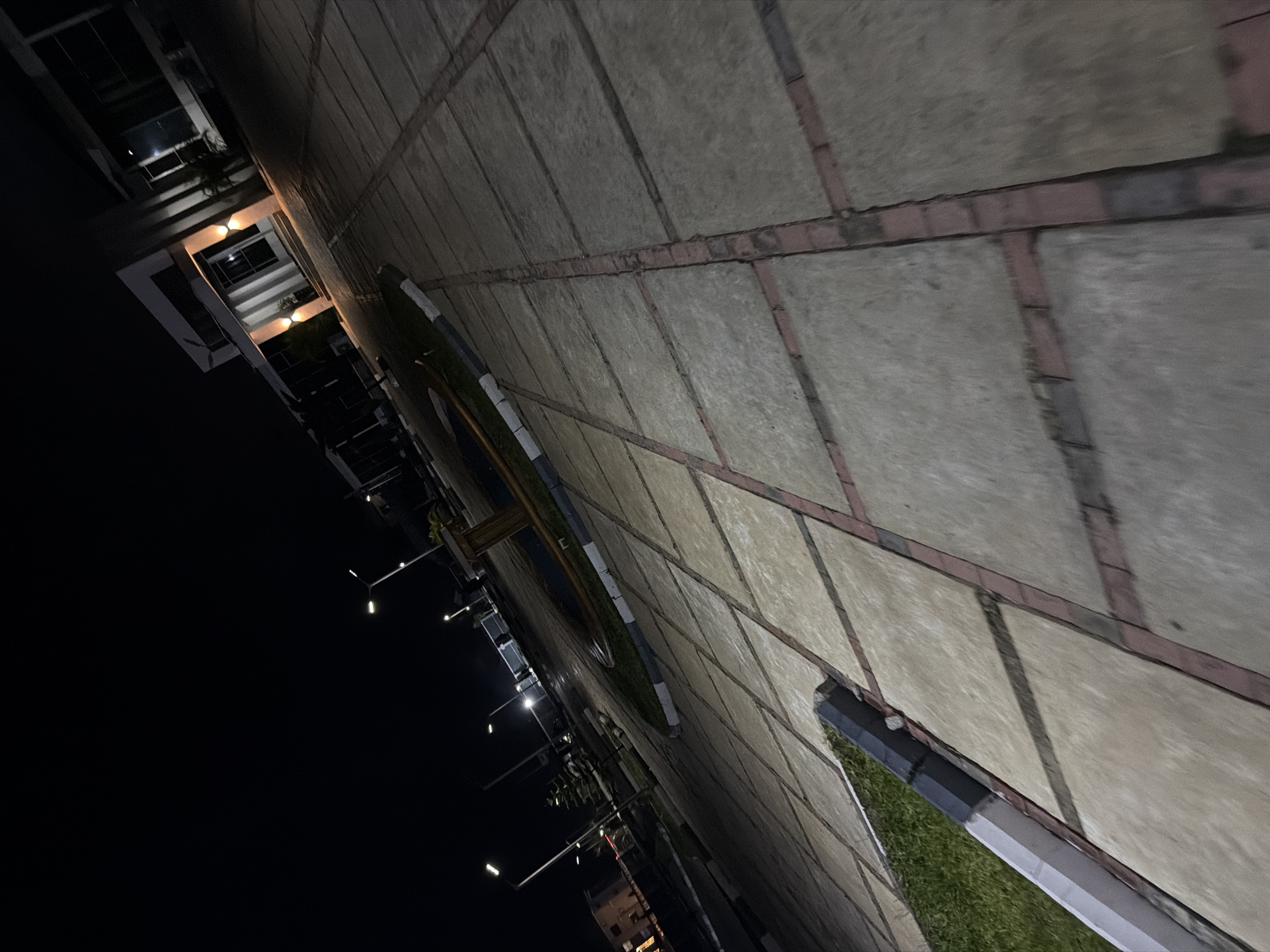 The image depicts an outdoor scene at night in Bukuru, Nigeria. A paved walkway, constructed with large, light-colored square tiles bordered by red bricks, dominates the foreground and midground. The walkway is angled diagonally from the bottom left to the top right of the frame. To the left of the walkway, a strip of green grass is visible, bordered by a concrete and dark grey segmented curb.
In the midground, a circular garden feature with some greenery is present, surrounded by a decorative border. Several modern streetlights with white lamps are spaced along the walkway, casting illumination on the paved area and the surrounding environment.
In the background, a modern building with large windows and a flat roof is illuminated. Two outdoor lights are visible on the facade of the building, casting a warm glow. The overall impression is that of a well-lit public space or commercial area in Bukuru at night. No people or animals are visible in the frame.