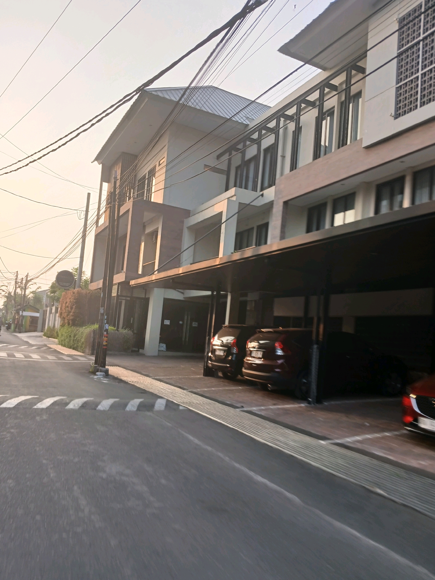 The image depicts a street scene in Depok, Indonesia, characterized by a modern building with a covered parking area and overhead utility wires. The building, likely a multi-story residence or commercial establishment, features a clean, contemporary design with large windows and a dark-colored, cantilevered roof over the parking area. Parked beneath the overhang are several vehicles, including a red SUV and a darker SUV. The street in the foreground is asphalt, marked with white pedestrian crossing lines and a speed bump. The environment appears to be a residential or mixed-use urban area, with hints of other buildings and lush greenery to the left of the main structure. The lighting suggests it is either early morning or late afternoon, with the sky appearing bright and hazy. There are no discernible people in the frame, and the overall atmosphere is still and quiet.