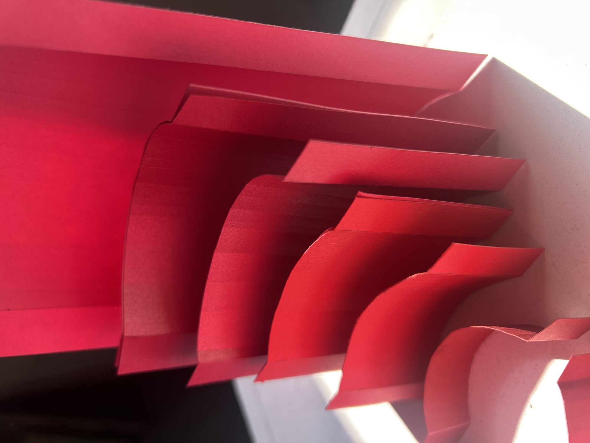 The image shows a close-up view of a red accordion file folder. The folder is positioned at an angle, with the individual sections fanning out from left to right. The sections are made of red paper or cardstock, and the edges of each section are clearly defined. The background is blurred, with areas of white and dark space. The lighting is uneven, casting shadows and highlights on the folder. The city and location are unknown.