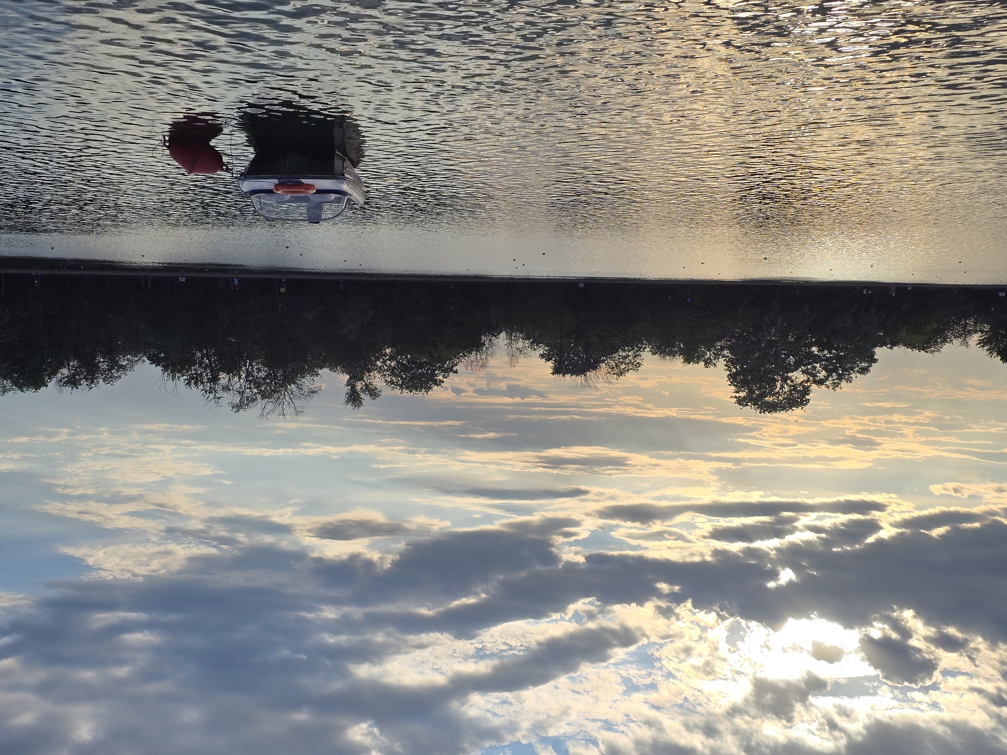 The image shows a body of water, possibly a lake or reservoir, with a small boat or buoy visible. The sky above is filled with clouds, with sunlight breaking through in some areas. A line of trees or vegetation is visible along the horizon. The image appears to be upside down. The location is Unknown, Unknown.