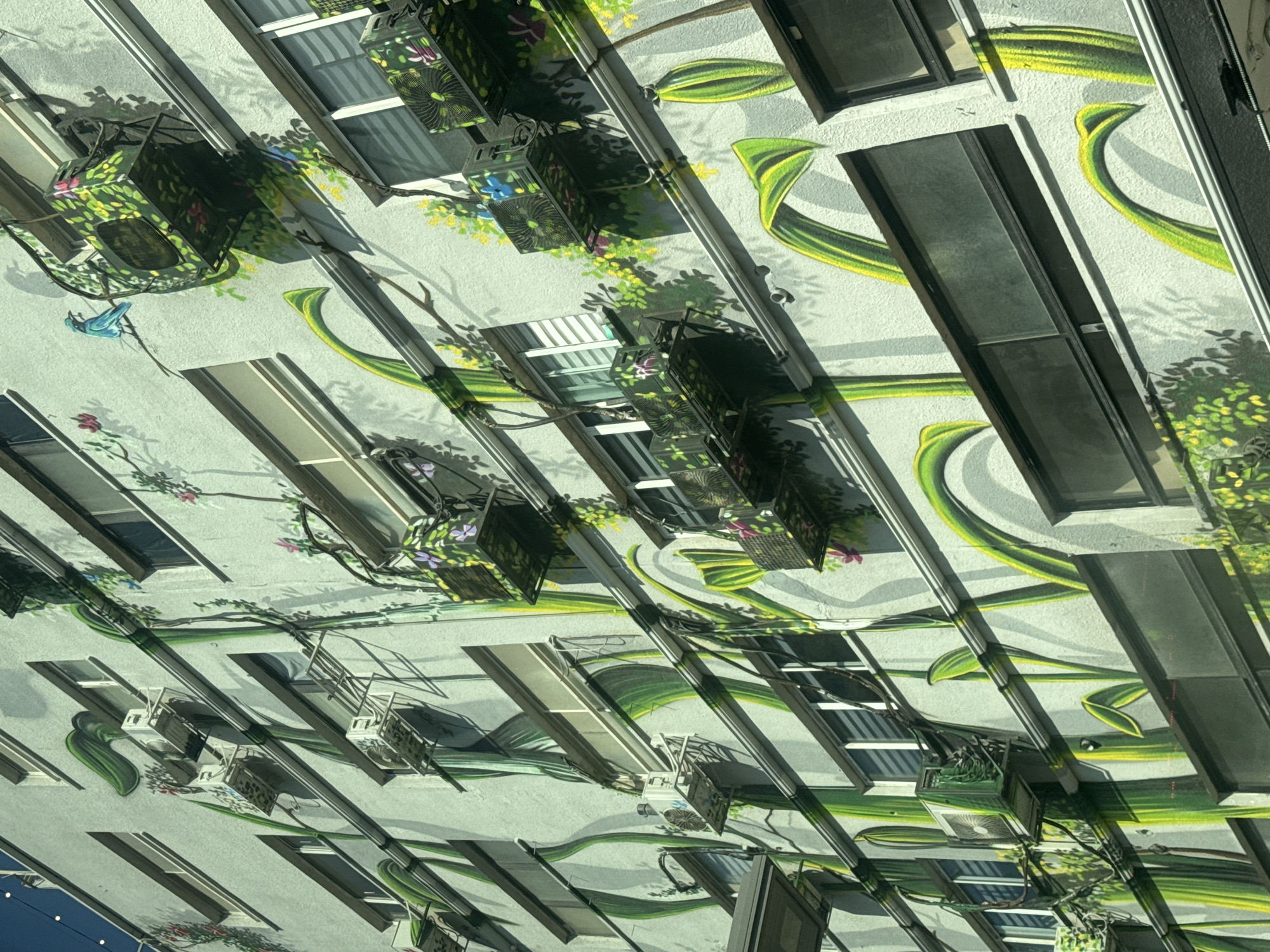 This image captures a section of a multi-story building facade in Tel Aviv, Israel, adorned with a vibrant mural. The artwork depicts stylized green plants, vines, and flowers intertwining with the building's architectural features. Several window units, air conditioning units, and pipes are visible, some of which have been integrated into or covered by the mural's design, creating a trompe-l'oeil effect. Specifically, some air conditioning units are decorated with greenery and flowers, blending them into the painted environment. A blue bird is perched on a branch as part of the mural. The building's exterior appears to be light-colored stucco, and the windows are mostly covered with blinds. The overall scene suggests an urban art installation designed to enhance the building's aesthetic.