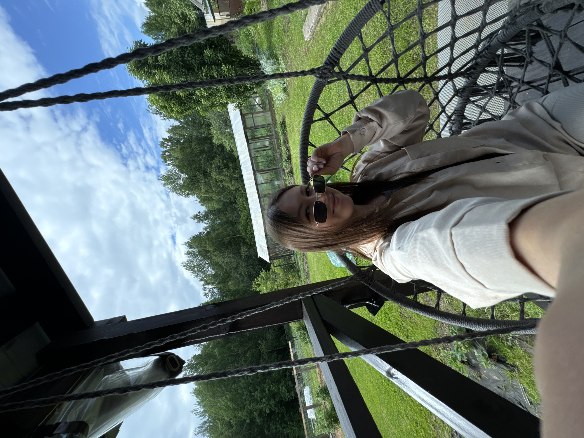 In the city of Ponazyrevo, Russia, a young woman with long brown hair is seated in a hanging chair on a wooden porch. She is wearing sunglasses and a light-colored collared shirt, and is touching her sunglasses with her right hand. The background shows a lush green garden with a greenhouse, trees, and a blue sky with clouds. The porch has dark wooden beams and supports, with thick ropes suspending the chair.
