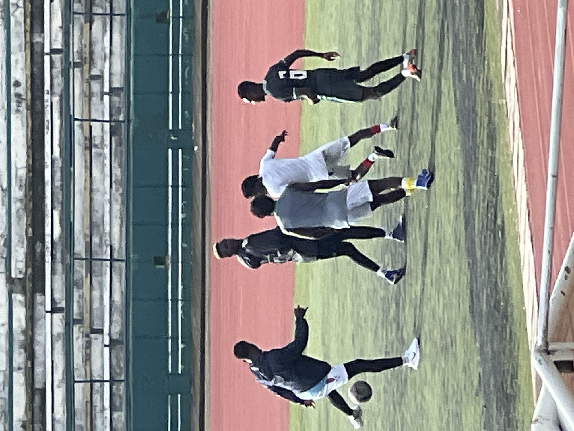 In Rafin Par, Nigeria, on a green turf soccer field, five young Black men are engaged in a soccer practice session. The men are wearing athletic attire, with one in a dark blue jacket and white shorts, another in a black shirt and shorts, a third in a grey sleeveless shirt and white shorts, a fourth in a white t-shirt and white shorts with red accents on his socks, and the fifth in a dark green shirt with the number "6" on the back and dark shorts. One player is actively kicking a soccer ball. In the background, stadium seating with weathered white benches and a green barrier are visible. The lighting suggests it is daytime, with shadows cast on the field.
