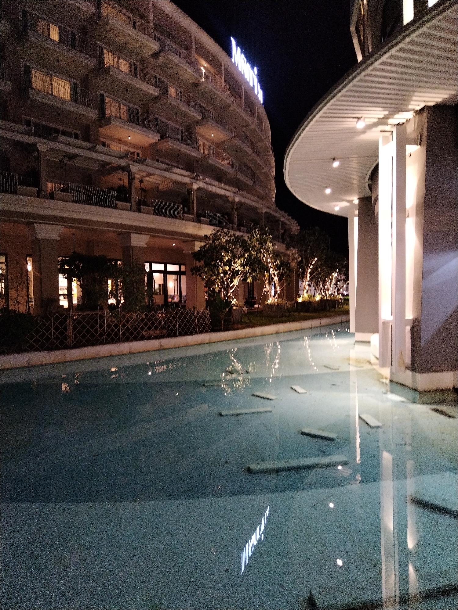 The image depicts a nighttime view of a resort in Patong, Thailand. On the left, a multi-story building with numerous balconies, illuminated from within by warm light, dominates the scene. The building's facade is a light color, and some balconies have decorative railings. To the right, a modern architectural element with a curved roof, supported by white pillars, is visible. This structure is illuminated by recessed lighting. In the foreground, a shallow, reflective pool or water feature occupies a significant portion of the frame. The surface of the water mirrors the lights from the building and the surrounding trees, creating a blurred, abstract pattern. Several rectangular stepping stones are submerged in the water. Trees with illuminated string lights are visible in the middle ground, between the building and the water feature. The overall atmosphere is tranquil and illuminated by artificial light against the dark night sky.