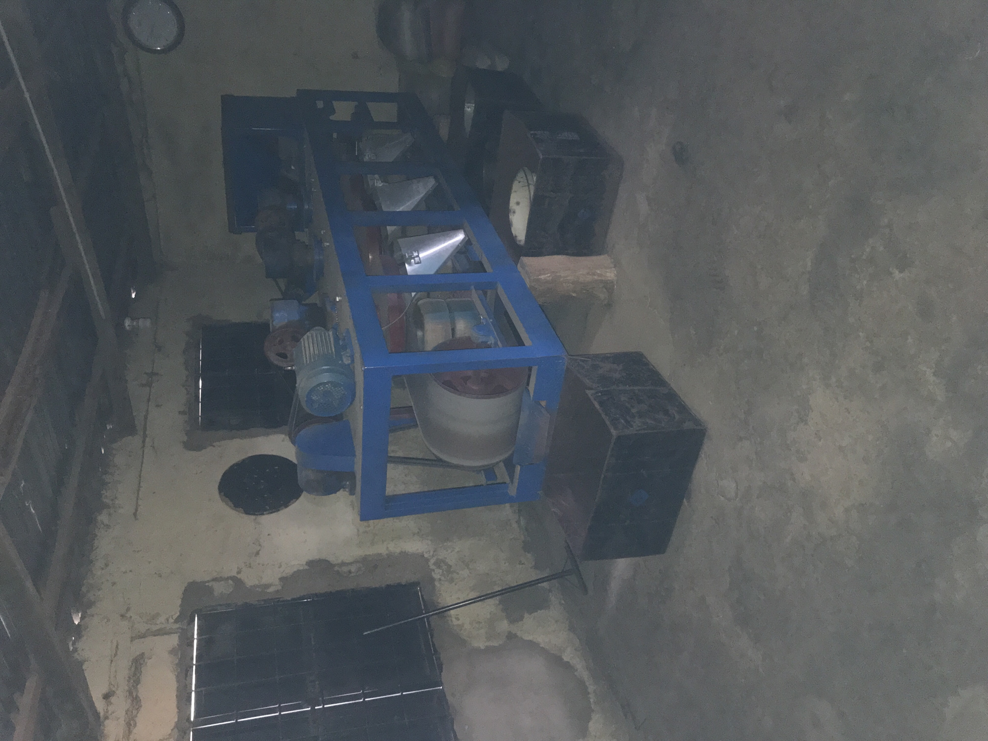 The image depicts the interior of a workshop or factory space, potentially located in the city of Kabong, Nigeria. The room has rough, unfinished walls, a corrugated metal ceiling, and a concrete floor. In the center of the room is a large, blue metal machine, likely industrial in nature, with various components including motors, belts, and a large roller. The machine is supported by a blue metal frame. In front of the machine are several black, open-top containers or bins, some resting on the floor and others raised slightly. A long-handled broom or rake leans against the wall near a barred window. A round clock is visible on the far wall. The lighting in the room is dim and appears to be artificial. There are no people visible in the image. The overall impression is of a functional, albeit somewhat rudimentary, industrial workspace.