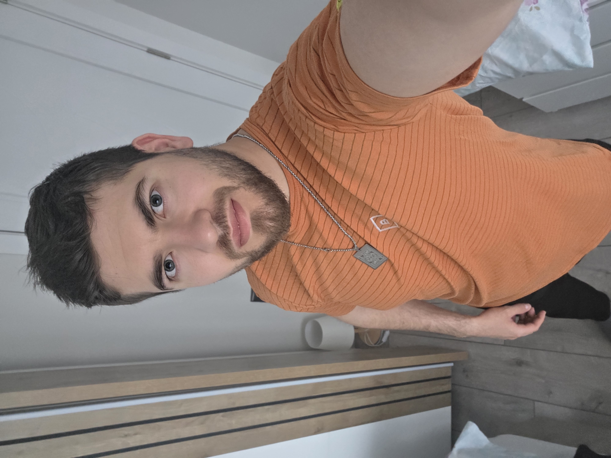 A man with dark hair, a beard, and blue eyes is facing the camera, wearing an orange shirt and a silver chain with a pendant. He is in an indoor setting, with a white and light wood-colored dresser behind him. A white cylindrical object, possibly a lamp base, sits on the dresser. A partially visible bed with white bedding is to the upper right of the man, and dark flooring is in the lower right. There is no evidence that suggests the location is Yagodovo, Bulgaria.