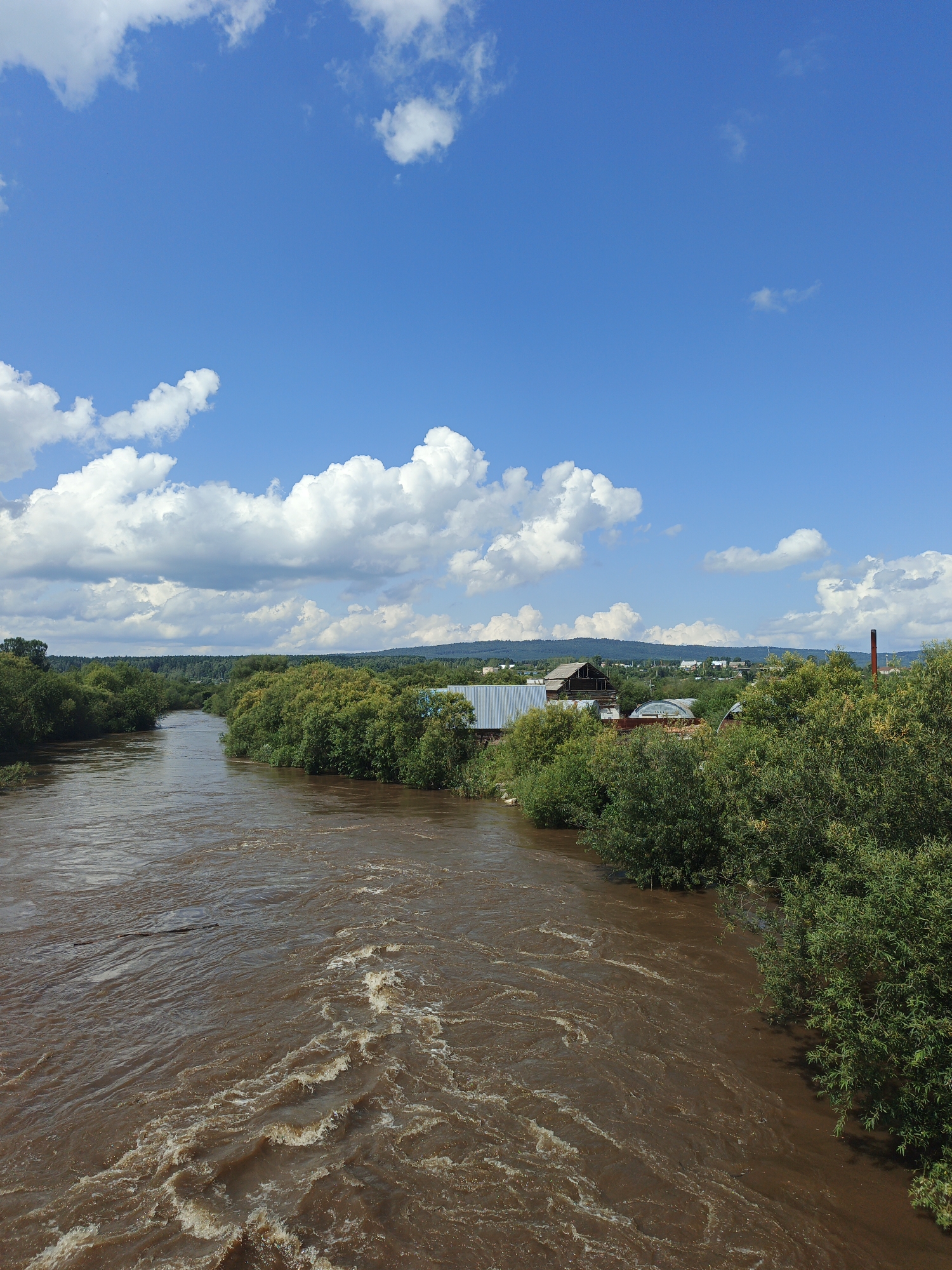The image depicts a wide, brown, muddy river flowing through a rural landscape. Dense green trees line the banks of the river, with a few scattered buildings visible in the background. The sky is a vibrant blue with scattered white, fluffy clouds. The scene appears to be a sunny day.
In the town of Medvedyovka, Russia, this image captures a view of the Belaya River. The river's muddy color suggests recent rainfall or sediment runoff. The dense foliage on the riverbanks indicates a natural, undeveloped area surrounding the settlement. The buildings in the background are typical of a rural Russian village, possibly including residential homes and outbuildings. The clear blue sky with clouds suggests fair weather, ideal for outdoor activities or agricultural work. The overall impression is one of a tranquil, natural setting with a touch of human habitation.