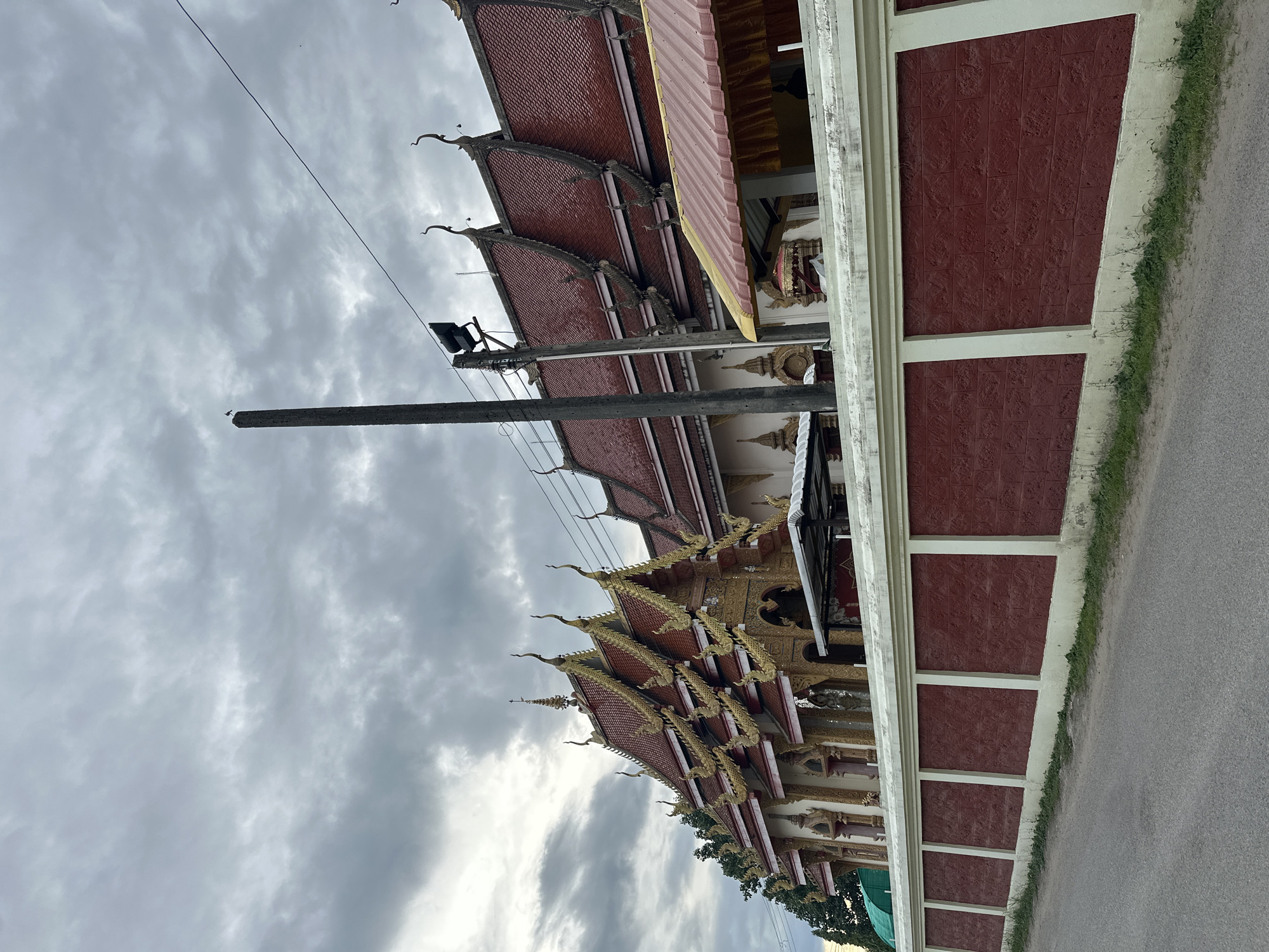 The image shows a temple in Ban Patan, Thailand, under a cloudy sky. The temple features a red tiled roof with ornate golden trim, and a tall, red brick wall with white accents separates it from the street. A concrete utility pole stands in front of the temple, with a bird perched on top. The sky is overcast with grey clouds. The scene is peaceful and somewhat subdued due to the weather.