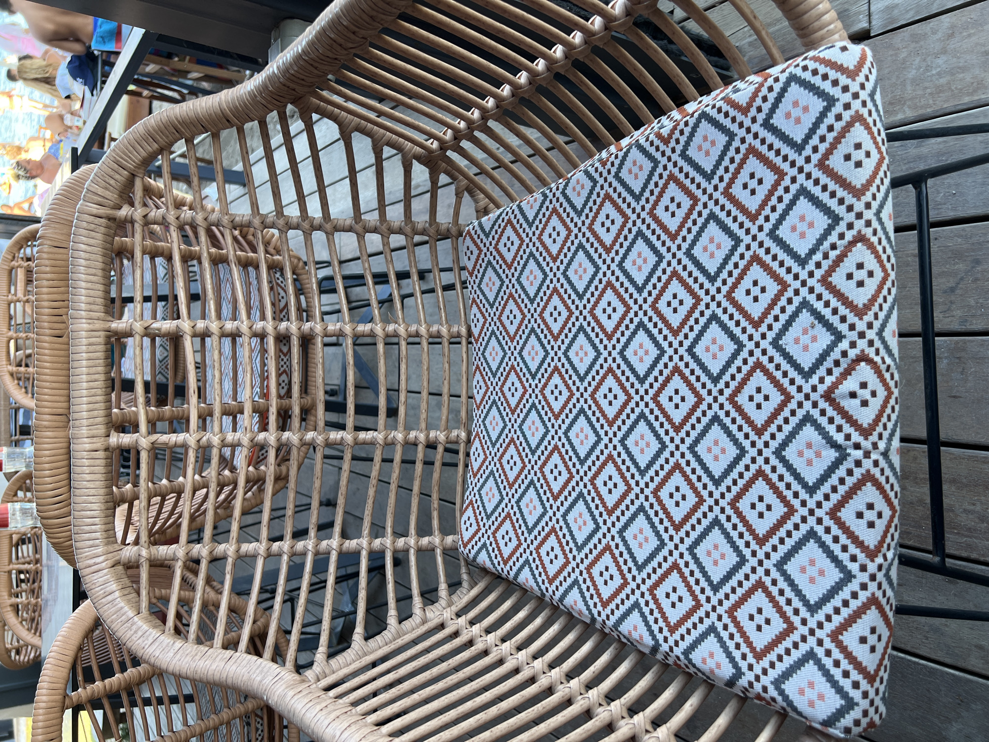 The image shows a close-up of a woven wicker chair with a patterned cushion. The cushion has a geometric design featuring diamonds in shades of white, brown, grey, and pink. The chair appears to be part of an outdoor seating area, possibly at a restaurant or cafe, with other similar chairs visible in the background. In the further background, people can be seen seated at tables near a body of water, suggesting a coastal or lakeside location. The overall setting appears to be a sunny, relaxed environment, consistent with a vacation spot. The location is identified as the city of Eretria Municipal Unit, Greece.