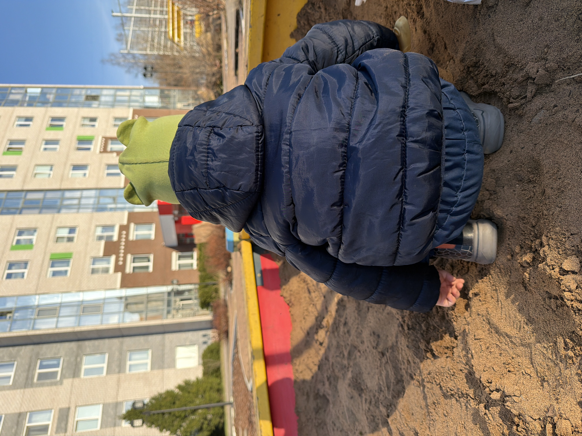 A child in a blue puffer jacket and green hat kneels in a sandbox. The sandbox has yellow and red edging. The child is touching the sand with their hands. Behind the sandbox, a multi-story building stands with various colored window frames. To the left, trees and bushes are visible. The image was captured on a sunny day. It cannot be confirmed from the visual data if the image was taken in Izhevsk, Russia.