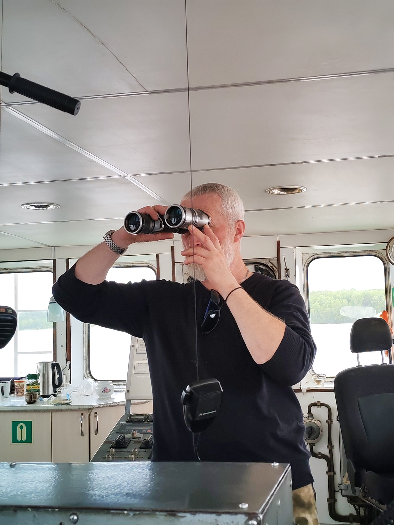 A man stands on a bridge of a vessel, possibly a ship or large boat, looking through binoculars. The bridge's interior has a utilitarian design, with a low ceiling and visible pipes and fixtures. The man has short gray hair and a beard and wears a black sweater. He is wearing a watch on his left wrist, and a bracelet on his right. Sunlight streams in through windows. The background landscape consists of water and a wooded shoreline. On a counter on the bridge a kettle, a jar labeled "Monarch", cups and other miscellaneous items are visible. A black chair is on the right side of the bridge. Given the mention of Turtsevo, Russia, the body of water visible through the windows is likely a part of the Volga river system or a connected waterway.