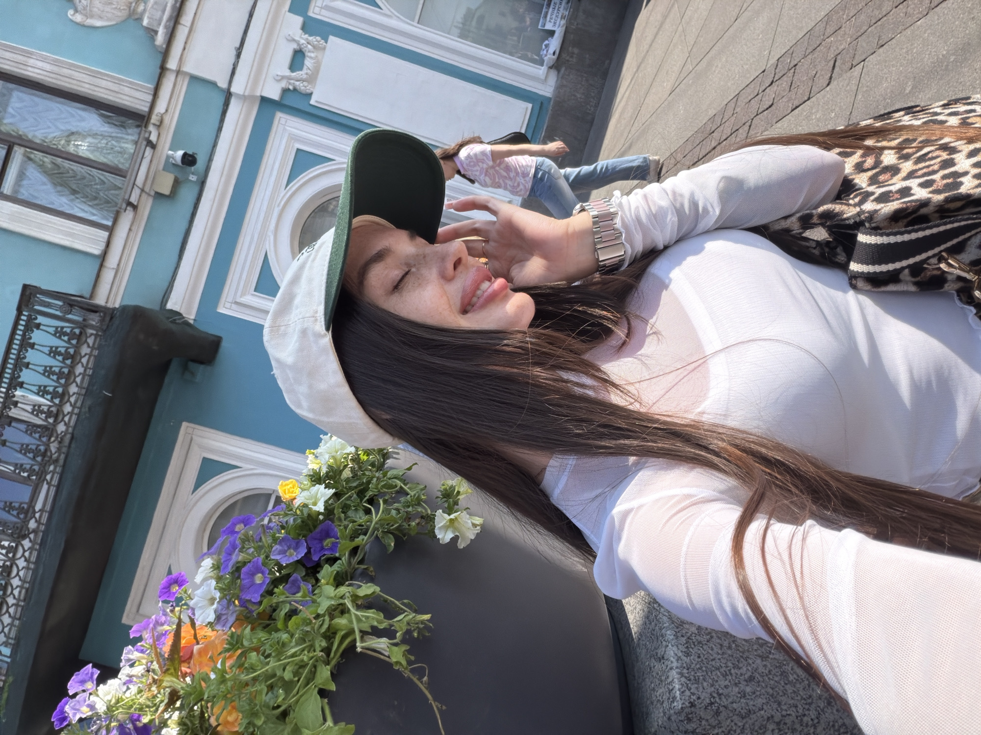 An outdoor, close-up, selfie-style photograph shows a young Caucasian woman with long, dark brown hair, wearing a white mesh top, a light-colored baseball cap with a green brim, and a silver wristwatch. She is smiling with her eyes closed and her tongue slightly extended, touching her face with her left hand. A portion of a large, dark gray planter filled with colorful flowers, including purple petunias, orange flowers, and white flowers, is visible to her left. A leopard-print handbag is slung over her right shoulder. In the background, a woman wearing a pink patterned shirt and blue jeans walks away from the camera. The scene is set against a backdrop of a light blue building with ornate white architectural details, windows, and a balcony. The ground appears to be paved with cobblestones or patterned tiles. The image is taken in Saint Petersburg, Russia, specifically along the Nevsky Prospekt, known for its vibrant street life and historic architecture.