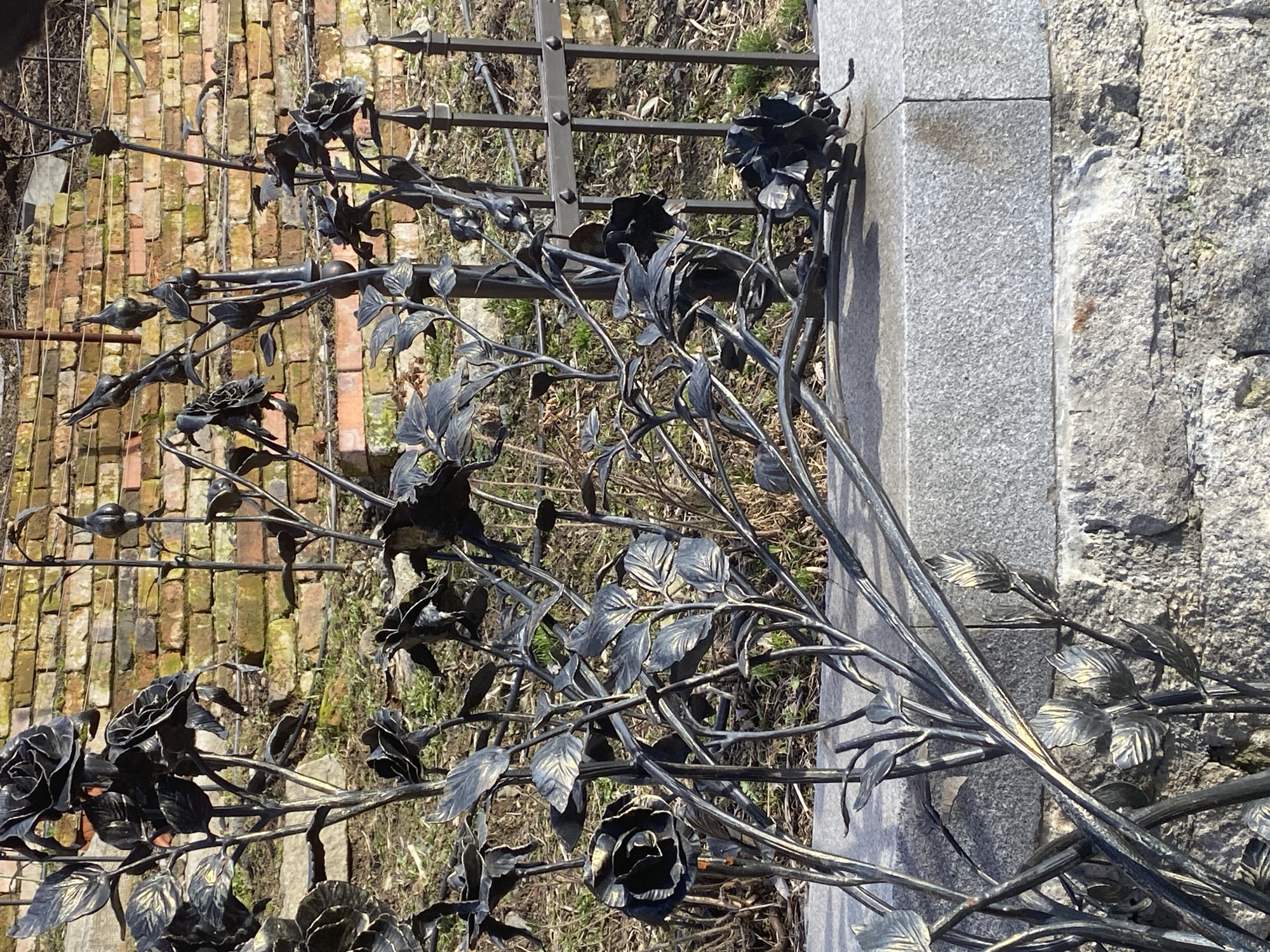 The image shows a wrought iron fence decorated with roses and leaves next to a tombstone. The background includes a brick wall and some sparse vegetation. The picture was taken at the Ivanovskoe Cemetery in Yekaterinburg, Russia.