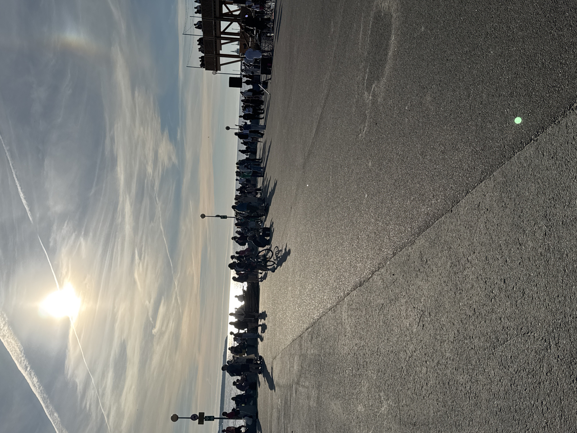 The image depicts a sunny day by the water, likely in Tallinn, Estonia, as suggested by the context of the scene which includes a promenade, people gathering, and a distinct wooden structure resembling an observation deck or stage. The sun is high in the sky, casting long shadows across the asphalt ground, indicating it's likely afternoon or late afternoon.
In the foreground, a wide expanse of asphalt pavement stretches out, with a subtle texture and a visible seam dividing different sections. The surface reflects the sunlight, creating a shimmering effect. To the left, a group of people are gathered along a low sea wall, some sitting and others standing, facing the water. The water itself is calm and reflects the bright sky, which is filled with wispy clouds and contrails.
Further into the scene, on the right side, a prominent wooden structure is visible. It has multiple levels and appears to be hosting a gathering of people on its upper platform, observing something below or in the distance. Below this structure, a band or musical setup is visible with equipment and individuals attending to it, suggesting a performance or event.
A diverse group of people are scattered throughout the scene. Some are casually strolling, while others are engaged in conversation or watching the proceedings. There are individuals with bicycles, and some appear to be children among the adults. The overall atmosphere appears to be relaxed and social, with a sense of community gathering.
The sky features a bright sun, with a faint rainbow-like arc visible on the right side, possibly a sun dog or atmospheric phenomenon. The light is diffused by the clouds, creating a soft, bright illumination. The environment suggests an outdoor public space, likely a waterfront area or promenade, popular for leisure activities and public events.