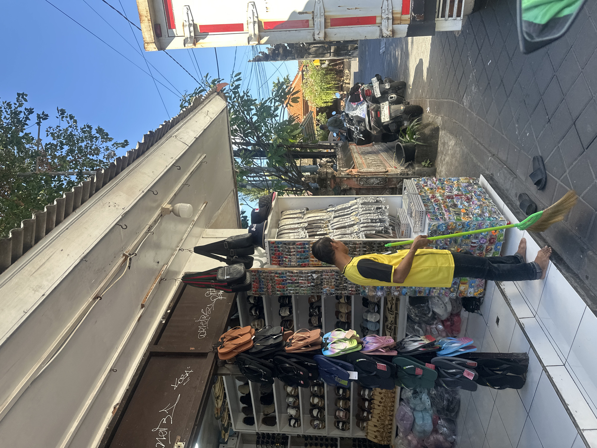 A street vendor in Kuta, Indonesia, is sweeping the pavement in front of his shop. The shop, which sells footwear and accessories, is packed with sandals and flip-flops displayed on shelves and hanging racks. The vendor, a man wearing a yellow t-shirt and dark pants, is using a green broom to clean the area. Behind the shop, a white truck is parked, and motorbikes are visible on the street. The scene is set against a clear blue sky and typical Indonesian architecture, with lush greenery and a tiled pathway.