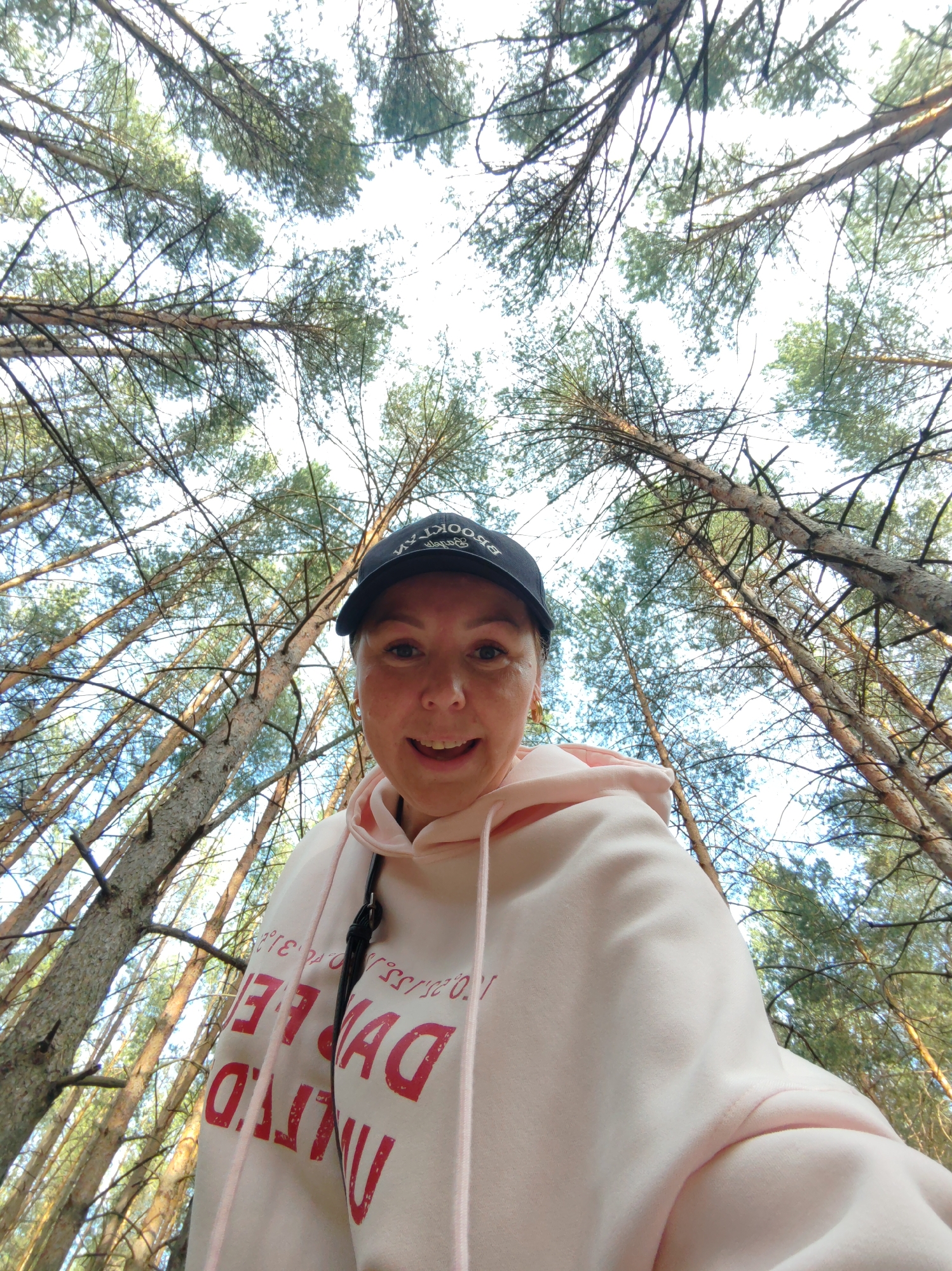 The image is a selfie taken by a woman in what appears to be a pine forest. She is wearing a light pink hoodie and a black baseball cap with the word "BROOKLYN" on it. Her hair is in a braid. The camera is pointed upwards, capturing the tops of the tall pine trees against a bright sky. The location is in a pine forest in the city of Chaikovsky, Russia. The woman is smiling.