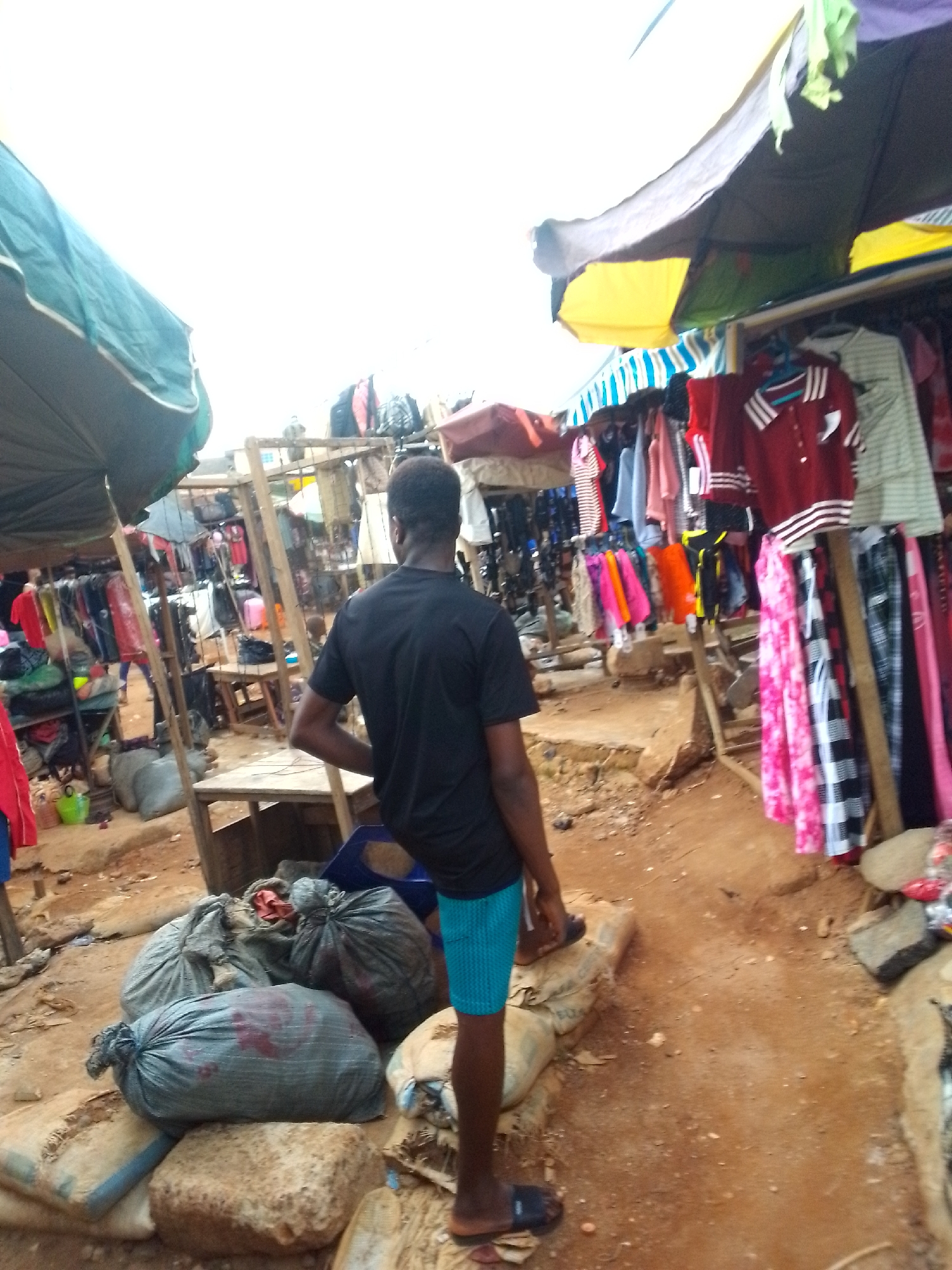 The image shows an outdoor market scene in Orlu, Nigeria. A man stands facing away from the camera, wearing a black shirt and blue shorts. He is standing on rocks or a raised surface. There are several large, tied bags on the ground near him. Clothes are displayed for sale on racks under canopies on both sides of a dirt path. There are wooden tables and stalls visible in the background. The area appears crowded with merchandise and structures.