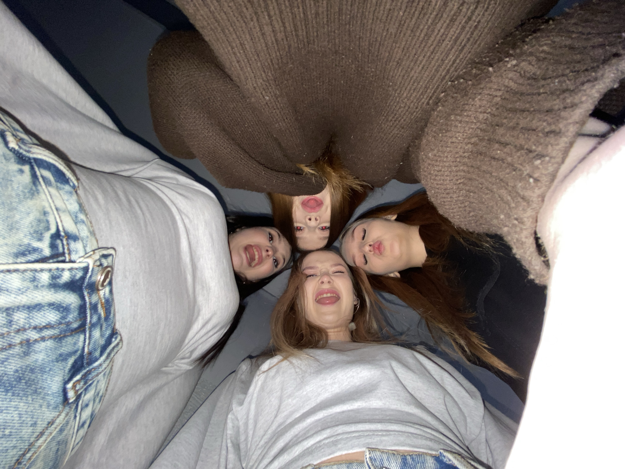 Four young women are huddled close together, posing for a photo taken from a low angle looking up. They are indoors, likely in a private space, based on their casual attire.
The woman closest to the camera is wearing a light gray sweatshirt and jeans. She has light brown hair and is making a silly face with her tongue sticking out. To the left is a woman with dark hair wearing a light gray sweatshirt with acid wash jeans and she is smiling.
Above the first woman is a third woman with auburn hair who is attempting to make a kissy face. A fourth woman, with blonde hair, is at the very top of the frame, appearing upside-down and pouting slightly. She is wearing a large, brown sweater.
The background is indistinct, but suggests the presence of a wall and possibly furniture. The overall lighting is dim and warm.
There is no indication of the city of Chaikovsky, Russia in the image.