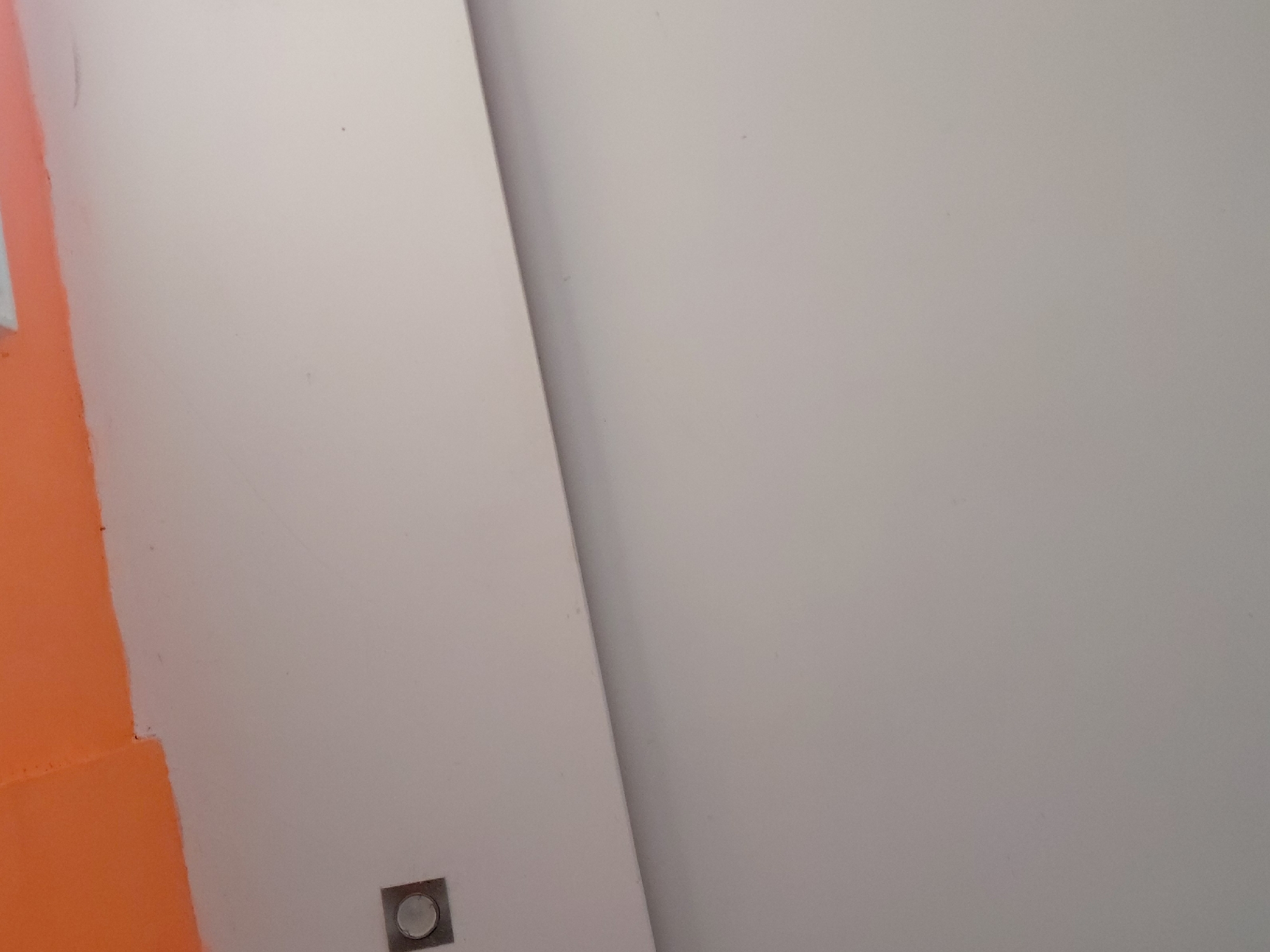 The image presents an abstract, close-up view of two surfaces with contrasting colors, divided by a diagonal line. The predominant area, covering the right and central parts of the frame, is a smooth, off-white or light gray surface. To the left, a distinct, vibrant orange surface is visible. A slightly irregular diagonal line separates these two color fields, running from the upper left to the lower right. In the bottom-left quadrant of the off-white section, a small, square-shaped metallic or dark gray fixture is visible, featuring a lighter circular element in its center, possibly a button or a rivet. The surfaces appear to be painted walls or panels, suggesting an interior setting. There are no people or discernible activities depicted. No text is visible. The lighting is even, and the scene emphasizes color blocking and simple geometric division.