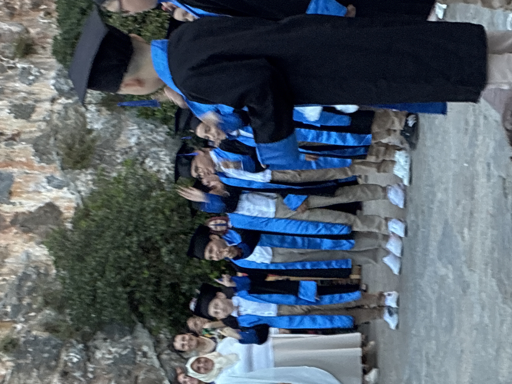 This image depicts an outdoor graduation ceremony or group photo taking place in Alanya, Turkey. A line of approximately seven individuals, likely graduates, are prominently featured, standing in profile and facing towards the right side of the frame. Each graduate is attired in a classic black academic gown and cap, strikingly accented with bright blue fabric on the sleeves and possibly as part of the stole or lining. Light-colored trousers or skirts and shoes are visible beneath their gowns.
The setting is characterized by a natural, rocky backdrop, suggesting a hillside or a historical site with a rugged stone wall texture. Green shrubs and small trees are scattered among the rocks, providing natural elements to the scene. The ground beneath the graduates appears to be a light-colored, possibly paved or stone surface. In the foreground and to the lower left, several other individuals, some dressed in casual wear or white garments, are visible, likely family members or attendees, observing the event. The lighting indicates it is daytime, possibly late afternoon, with soft, even light illuminating the scene. The overall mood is one of ceremony and celebration. No visible text is present in the image.
