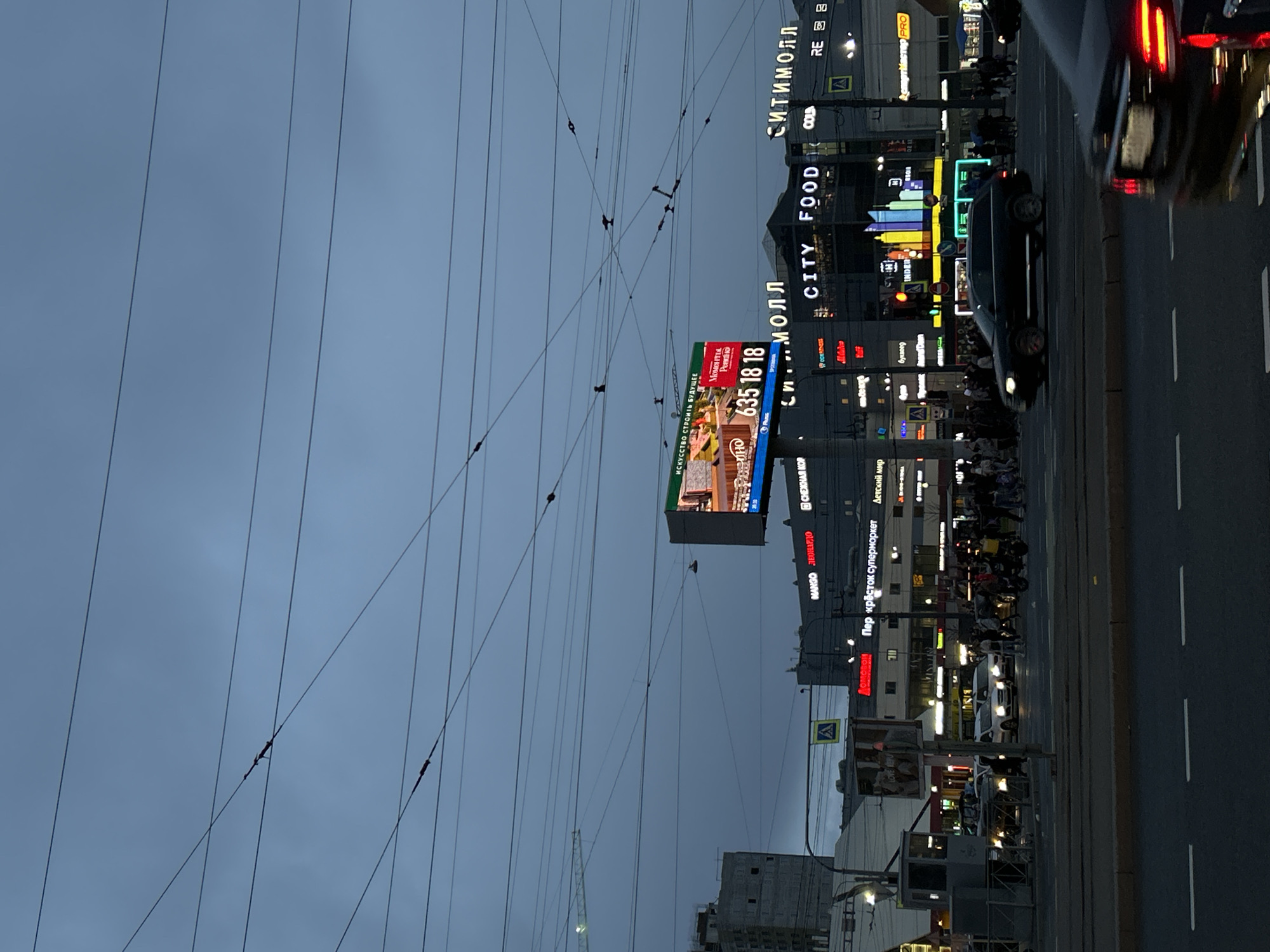 An urban street scene in Saint Petersburg, Russia, is captured at dusk or early evening. The sky is a deep blue-grey, with a complex network of overhead power lines crisscrossing prominently across the upper and left parts of the image.
The scene features a busy street with vehicles and pedestrians. A multi-lane road runs along the right side, showing cars in motion, indicated by the bright red taillights of a vehicle in the foreground and another dark car further down. Along the right side of the street, commercial buildings line the area, illuminated by a multitude of neon and backlit signs. Pedestrians can be seen walking on the sidewalks.
A large, brightly lit billboard stands out in the mid-ground, displaying an advertisement with images of people. Text on this billboard reads: "ИСКУССТВО СТРОИТЬ БУДУЩЕЕ" (Art of Building the Future), "Моменты.", "Романо", and a phone number "635 18 18".
Numerous other signs for various businesses are visible on the buildings, including "СИТИМОЛЛ" (CITYMALL), "CITY FOOD", "COLA", "RE", "MANGO", "ЛЕОНАРДО" (LEONARDO), "Пер крёсток супермаркет" (Perekrestok Supermarket), "Детский мир" (Detsky Mir), "буквоед" (Bukvoed), "INDER", and "рrмастер PRO". The abundance of illuminated signs and the dark sky confirm it is either late evening or night, creating a vibrant city atmosphere.
