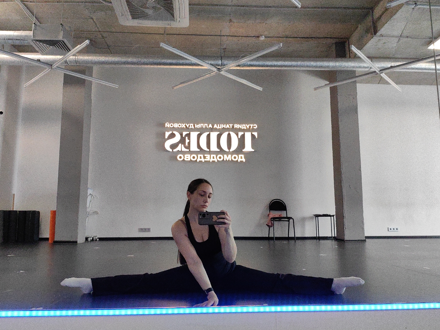 A young woman is captured in a selfie taken in a large mirror, showcasing her flexibility in a full front split. She wears a black top and black pants, with white socks, and holds a smartphone to take the picture. Her expression is neutral and focused. The setting appears to be an indoor dance or fitness studio, characterized by a dark floor, white walls, and an industrial-style exposed concrete ceiling with visible ventilation ducts and modern, star-shaped light fixtures. Along the bottom edge of the mirror, a strip of blue LED lighting provides a distinct glow.
In the background, visible through the mirror's reflection, is a large, illuminated sign on the white wall. The main text, clearly decipherable when un-mirrored, reads "TODES". Directly below this, also un-mirrored, is "ДОМОДЕДОВО" (Domodedovo), indicating the location. A smaller line of stylized text, which is harder to precisely un-mirror, appears above "TODES". Other objects reflected in the background include a black chair with a light-colored item on it, a small black table, and some exercise equipment (an orange foam roller and black stacked items) near the left wall.