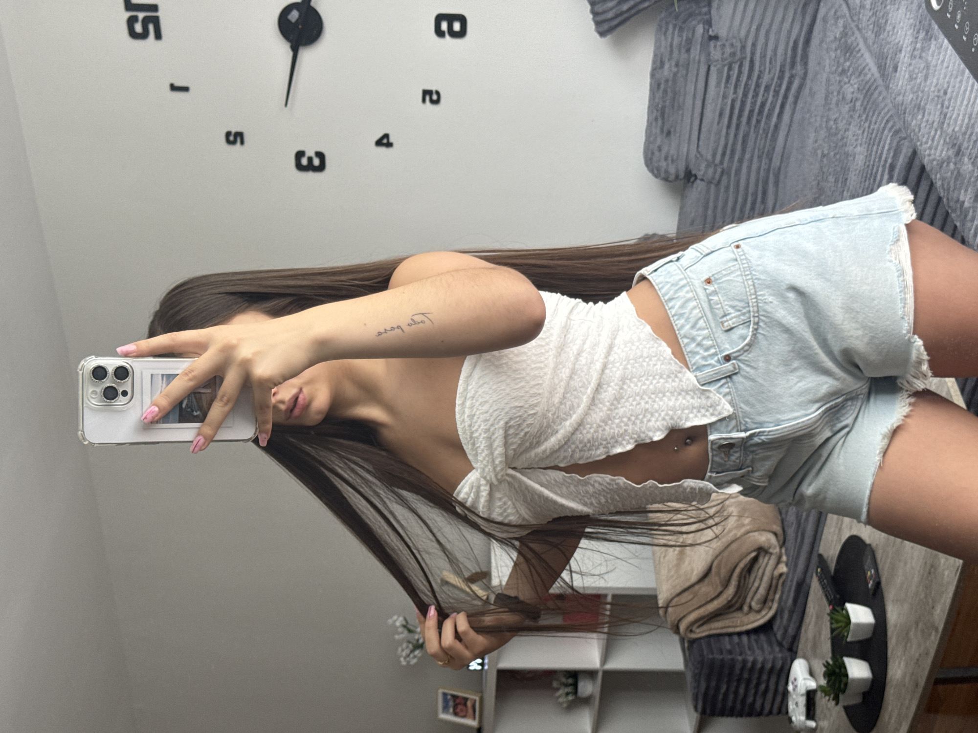 The image shows a young woman taking a selfie in a mirror. She is wearing a white textured crop top and light blue denim shorts. Her long, dark hair is down and falls around her shoulders. She has a belly button piercing and a small tattoo on her left forearm that appears to read "toca boca." She is holding a white smartphone with a clear case, which is reflecting an image of what looks like a horse.
The setting appears to be indoors, possibly a bedroom or living area. In the background, a large wall clock with black numbers and hands is visible. A grey, textured sofa or chair is also present. A blurred image of shelves with decorative items and a folded blanket adds to the domestic feel of the scene.
The lighting suggests it is daytime, but no specific time of day can be determined from the clock's hands. The woman's expression is neutral as she focuses on taking the photo. The overall mood is casual and personal. The location context of New York, USA, is provided, but there are no specific visual cues in the image that directly confirm this location.