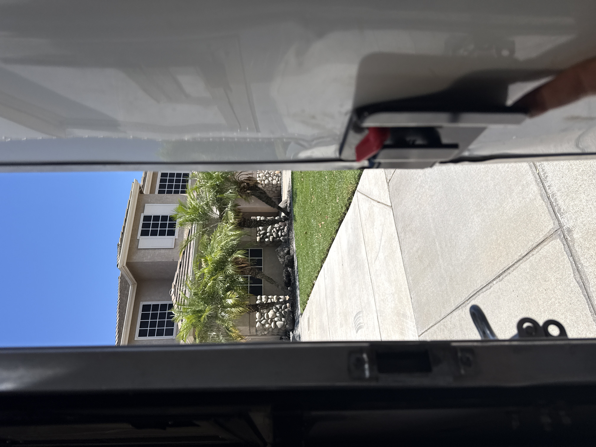 The image, taken on a sunny day in Ripon, California, shows a view from inside an open doorway, likely of a vehicle or storage unit. The perspective looks outward towards a two-story house with a light-colored stucco exterior, white-trimmed windows, and a shingled roof. The house is surrounded by a neatly manicured green lawn and several palm trees. In the foreground, a concrete sidewalk leads away from the viewer towards the house. A black door frame is visible on the left, and the white side of what appears to be a vehicle or storage unit dominates the right side of the frame. A red latch mechanism is visible on the white surface. The sky is clear blue.