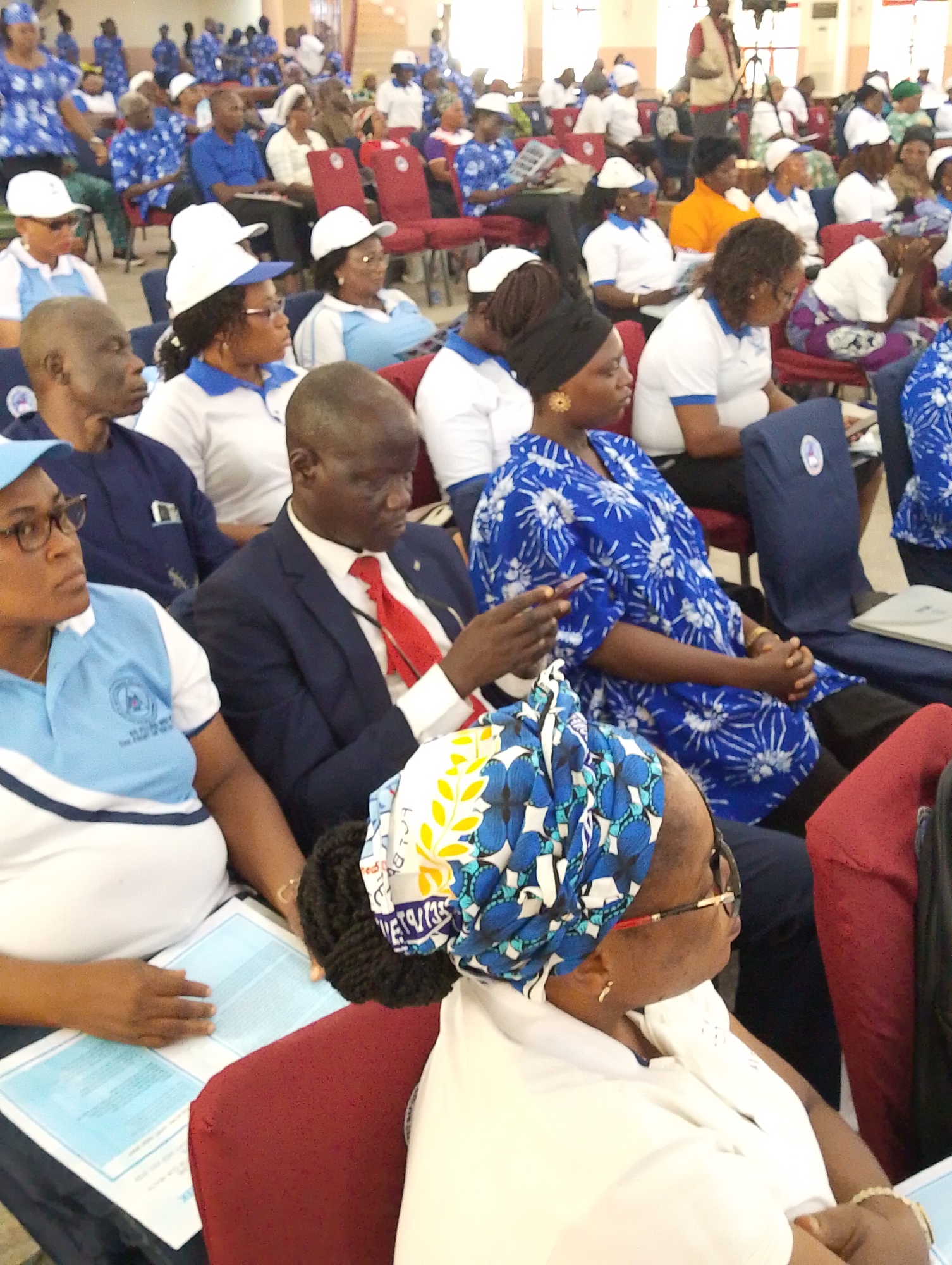 A large indoor gathering is captured from within the audience, showing numerous people seated in rows of red and dark blue chairs. The scene suggests a conference, meeting, or official event in Kubwa, Nigeria.
The attendees are predominantly adults, dressed in a mix of formal and semi-formal attire. A significant number of individuals, particularly women, are wearing blue and white patterned tops or uniform-like shirts, often paired with white baseball caps or headscarves. One woman in the immediate foreground wears a distinct blue, white, and yellow patterned headwrap. Many men are seen in blue polo shirts, while one prominent man in the mid-foreground wears a dark suit with a bright red tie.
People are generally attentive, looking towards the front of the room, though some are seen interacting with papers or mobile phones. In the bottom left, a woman in a blue and white top is looking at a light blue document laid out on a table in front of her. Another woman in the deep foreground is also looking at documents.
In the background, more attendees fill the hall, and a person standing near what appears to be a tripod with camera equipment suggests media coverage or presentation setup. The overall atmosphere appears focused and organized. The lighting is bright and even, consistent with an indoor daytime event. No specific legible text from the documents or clothing is clearly identifiable beyond general patterns and logos.
