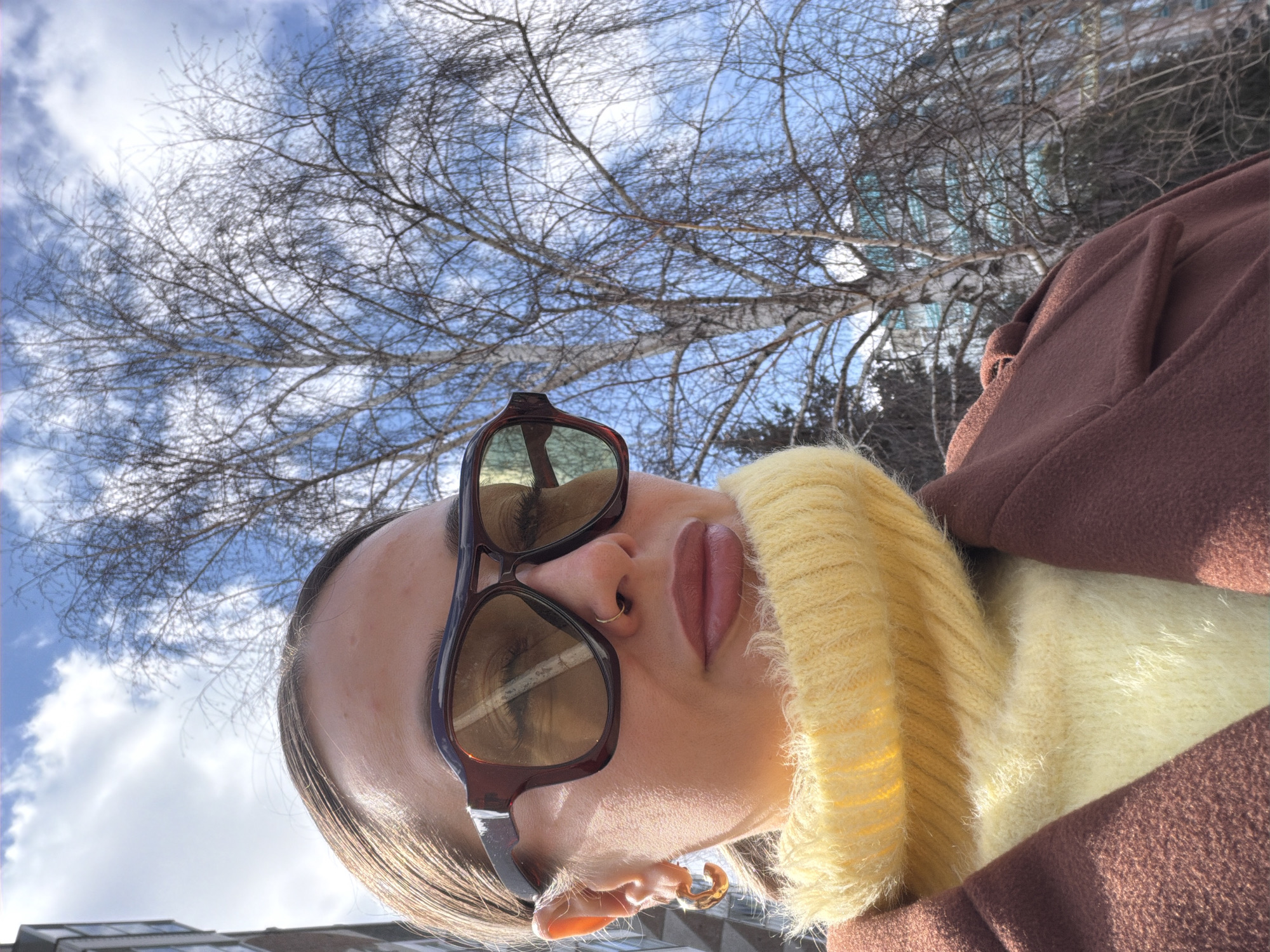 A low-angle outdoor shot features a person looking upwards, their head tilted back, against a bright blue sky with scattered white clouds. The individual wears a fuzzy, high-neck yellow sweater and a brown coat. Large, dark-rimmed sunglasses with amber lenses cover their eyes, reflecting the intricate network of bare tree branches. The person's lips are painted a dark rose hue, and they have a small gold hoop nose ring and a matching C-shaped earring. The background is dominated by a dense array of leafless deciduous tree branches, possibly birch, suggesting late winter or early spring. In the upper left background, partially obscured by the trees, is a multi-storied urban building with numerous windows. Bright, natural sunlight illuminates the scene, indicating midday or early afternoon. The setting appears to be an urban green space or street in Krasnoyarsk, Russia. No text is visible in the image.