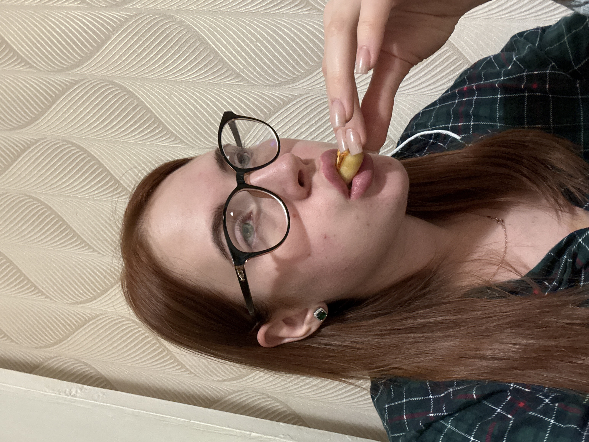 A woman with red hair, wearing glasses and a green and red plaid shirt, is holding a small, yellow food item (possibly a peanut or small piece of fruit) between her fingers and bringing it to her mouth. She is wearing a green earring on her left ear. The background is a beige wall with a wavy pattern. The location is not discernable from the image.