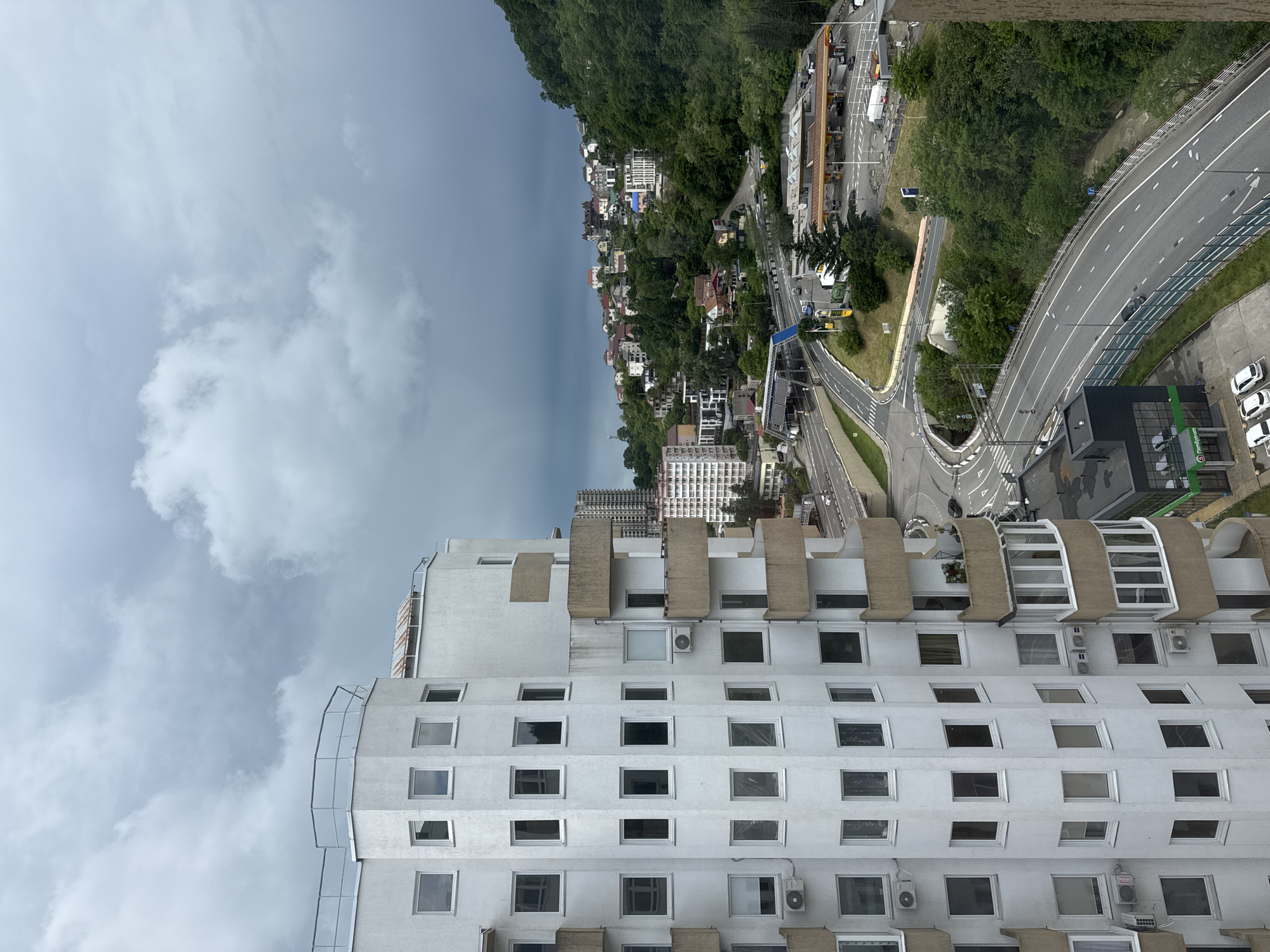 This image shows a view from a high vantage point of a building in Sochi, Russia, likely a hotel, overlooking the surrounding landscape. The building is white with multiple rows of square windows and protruding architectural features. In the background, there is a densely populated urban area situated along a hillside, with a road winding through it. On the right side of the frame, a modern highway is visible, with greenery separating it from the urban area. The sky is overcast with dark clouds.