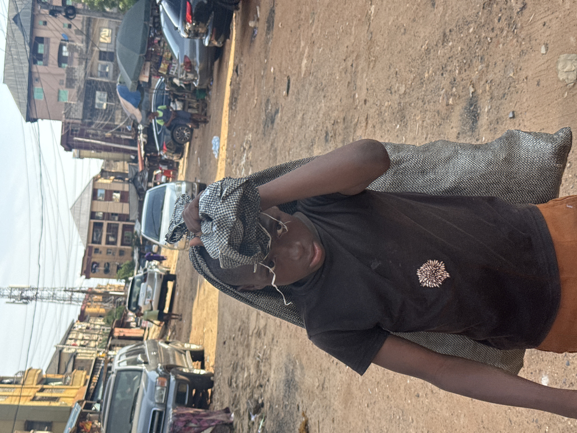 The image depicts a young person carrying a bundle on their head, which is covered by a grey and black patterned cloth. The individual wears a dark t-shirt with a white floral emblem on the left chest and orange shorts. The scene appears to be an outdoor marketplace in Onitsha, Nigeria. In the background, there are several buildings with multiple stories, some appearing to be shops or residences, and various vehicles, including cars and what might be a motorcycle. The ground is a dusty, sandy surface. The lighting suggests it is daytime, possibly late morning or early afternoon, with clear skies. The person's face shows a neutral expression. The overall impression is of daily life and commerce in a busy urban environment.