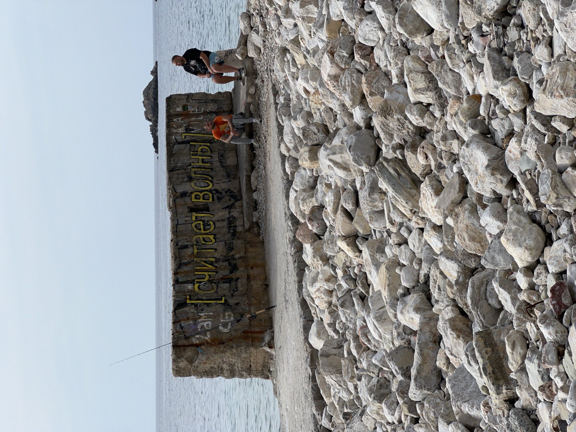 The image depicts a coastal scene at Perazića Do, Montenegro. The foreground is dominated by a large pile of gray rocks, providing a textured landscape. A concrete structure extends from the foreground towards the sea, featuring faded graffiti including what appears to be a yellow inscription in Cyrillic script. Two men are positioned on this structure. One man, wearing a dark t-shirt and shorts, is sitting on the edge of the structure, looking out towards the water. Another man in an orange shirt is sitting nearby, appearing to be engaged in fishing, as a fishing rod extends from the structure towards the water. The sea stretches out towards the horizon, and a rocky islet is visible in the distance. The sky is overcast.