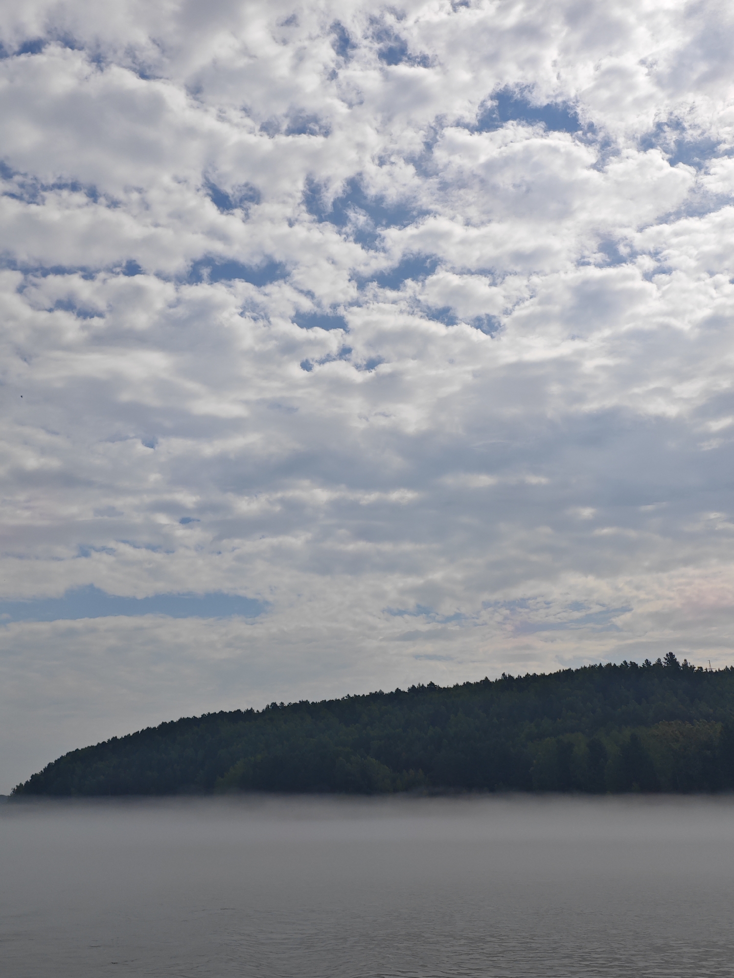 The image depicts a serene natural landscape. The foreground features a body of water with subtle ripples, possibly a river or lake. A thick layer of fog hovers just above the water's surface, obscuring the lower portion of the distant shore. The middle ground is dominated by a densely forested hillside, which rises steeply from the fog-shrouded water. The trees are a deep green, and the outline of the treeline forms a distinct curve against the sky. The sky above is filled with a complex pattern of white and gray clouds, with patches of blue visible in between. The overall impression is one of tranquility and natural beauty. The location is the city of Atamanovo, Russia.