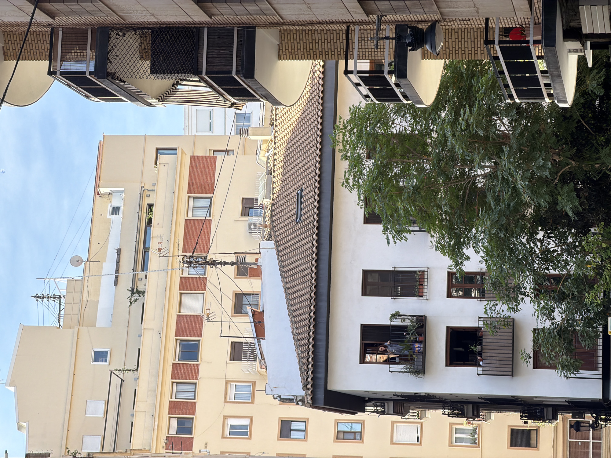 This urban photograph depicts a street scene in Valencia, Spain, characterized by a mix of traditional and modern multi-story residential buildings under a clear blue sky.
In the center-foreground, a prominent white building features a traditional terracotta tiled roof. On its top-floor balcony, a woman with dark hair, wearing a light-colored top, stands and looks directly out from the railing. Directly below this, a second balcony holds various potted plants, and a partial figure is visible behind its railing, also looking out. A large, leafy green tree partially obscures the lower sections of this white building and the immediate foreground.
To the left, a taller, light yellow/beige building rises, exhibiting multiple rectangular windows, some with closed blinds. Sections of red brickwork are visible between windows on its upper stories, and small balconies with dark railings project from some levels.
To the right, another multi-story building with a light brown brick facade features distinctive curved, concrete balconies with dark metal railings. A black, ornate street lamp is affixed to the underside of one of these balconies.
In the mid-ground and background, further light-colored buildings with various window configurations and rooftop structures are visible, including utility poles, an antennae array, and a satellite dish. Utility wires crisscross the sky between buildings. The scene is brightly lit by daylight, indicating sunny weather.