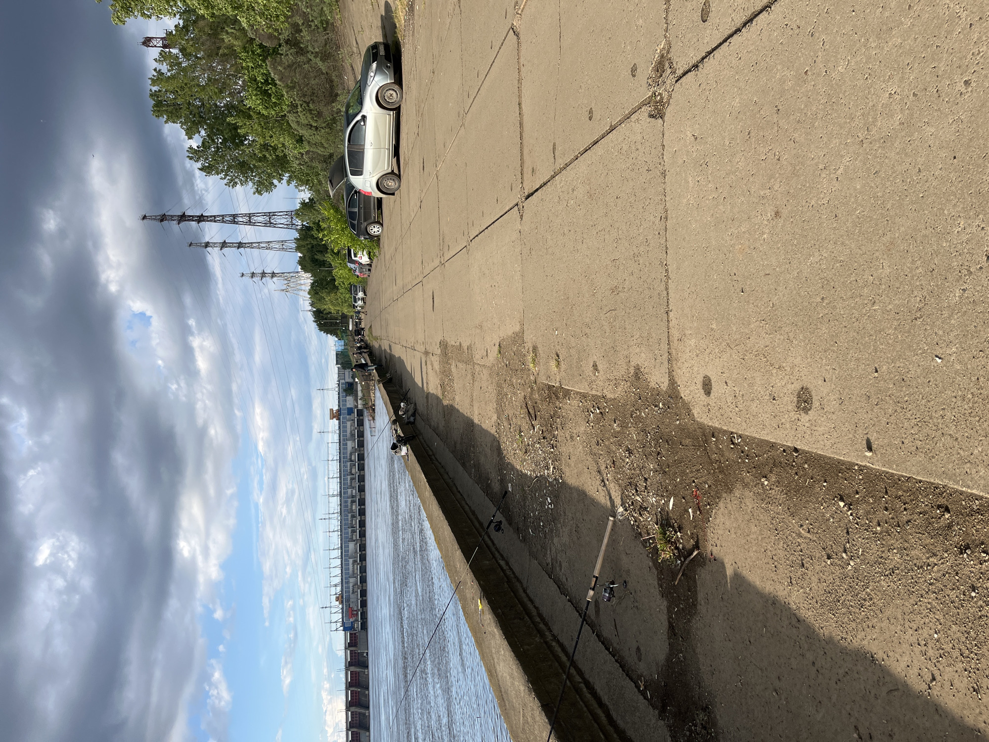 The image shows a concrete embankment in the city of Chaikovsky, Russia. On the right of the embankment is a sidewalk with parked cars. Along the edge of the embankment, several fishing rods are set up, indicating that people are fishing. In the background, there is a series of structures possibly related to power transmission or a dock. The sky is partly cloudy with power lines visible.