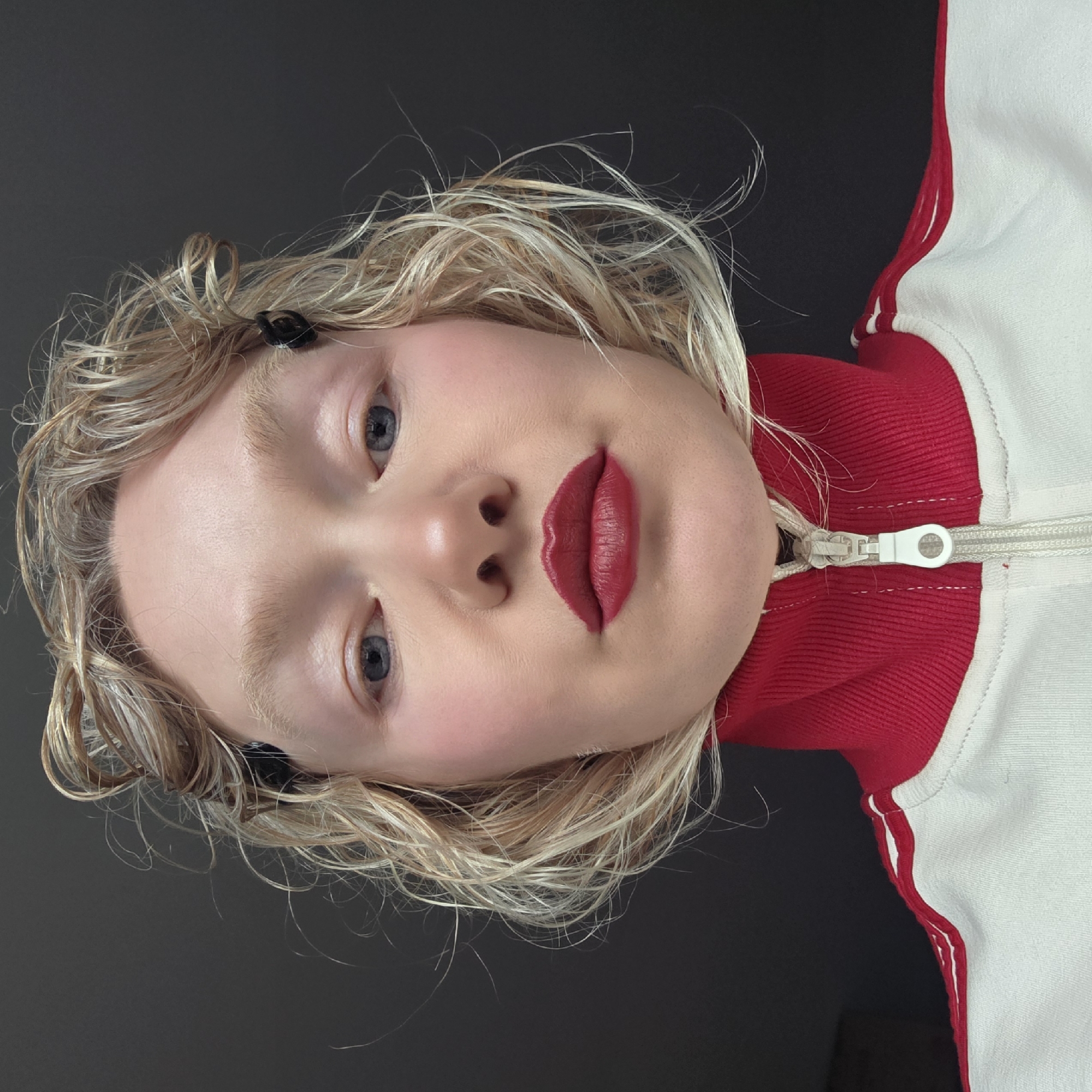 This close-up portrait features a young individual with fair skin, light blonde or strawberry blonde curly hair, and light blue eyes. The person's face is positioned at an angle, with a neutral and calm expression, and their gaze directed towards the viewer. Their lips are adorned with vibrant red lipstick, and a hint of pink blush is visible on their cheeks. Their eyebrows and eyelashes are very light, blending with their hair color. A small black hair clip is tucked into the hair above their right eye.
The subject is wearing a red and white athletic jacket, possibly a track jacket, featuring a ribbed red collar and a prominent white zipper running down the front. White panels or stripes are visible along the side of the jacket.
The background is a plain, solid dark grey or black, indicating an indoor setting or a studio shot with a simple backdrop. There are no other discernible objects, activities, or textual elements within the frame. The image does not contain any visual cues to determine the time of day, weather, or a specific geographical location, beyond the provided external context of Saint Petersburg, Russia.