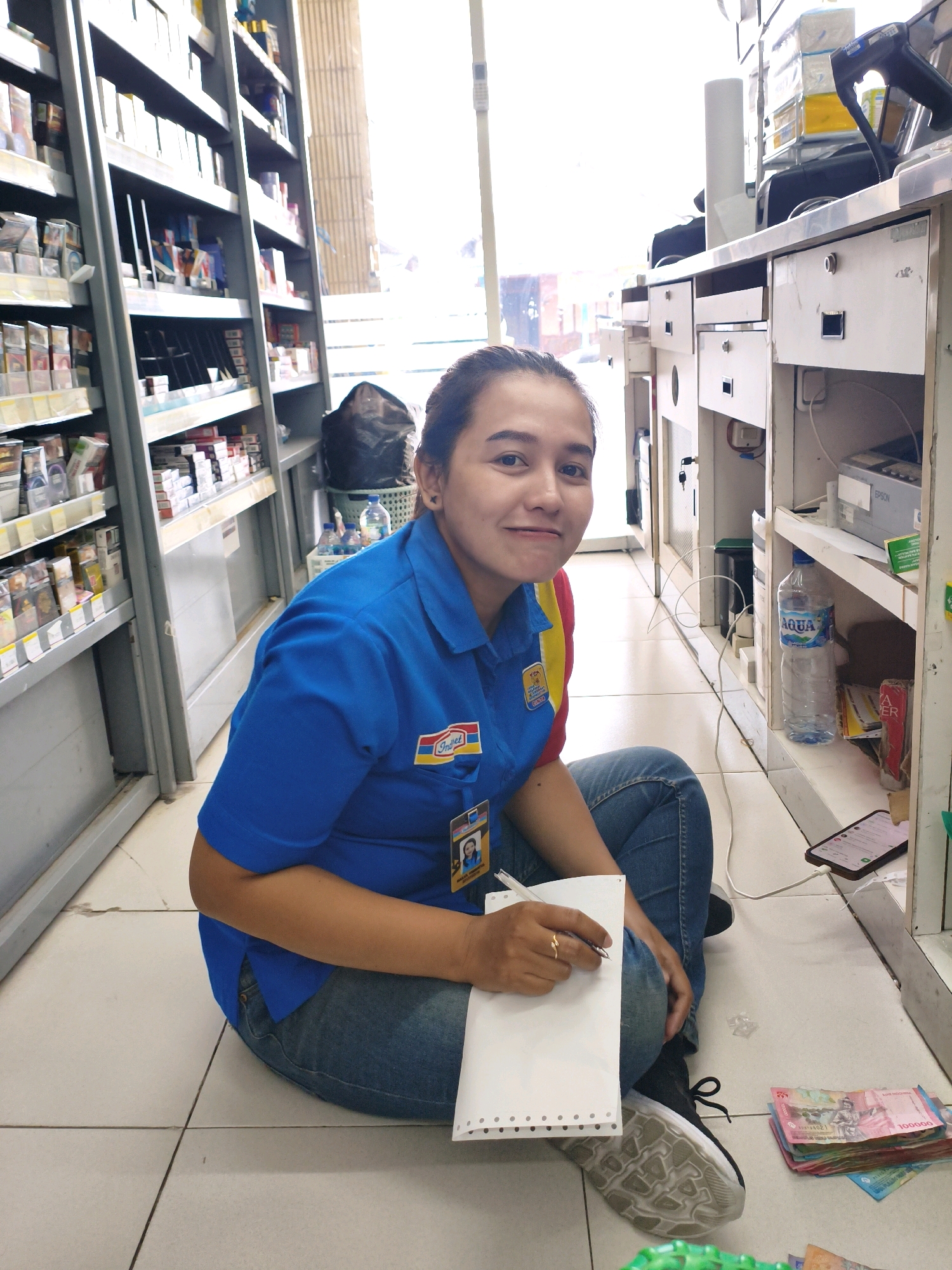 In a convenience store, likely in Sumberejo Kulon, Indonesia, a woman wearing a blue uniform with yellow and red trim is sitting on the tiled floor. She has a name tag visible on her uniform. She is holding a pen and writing on a white notepad. In front of her, a stack of Indonesian Rupiah bills is laid out on the floor. To her left, shelves stocked with various products, including cigarettes, are visible. To her right, a counter area with drawers, a printer, and a bottle of "AQUA" water are present. The overall environment suggests a retail setting.