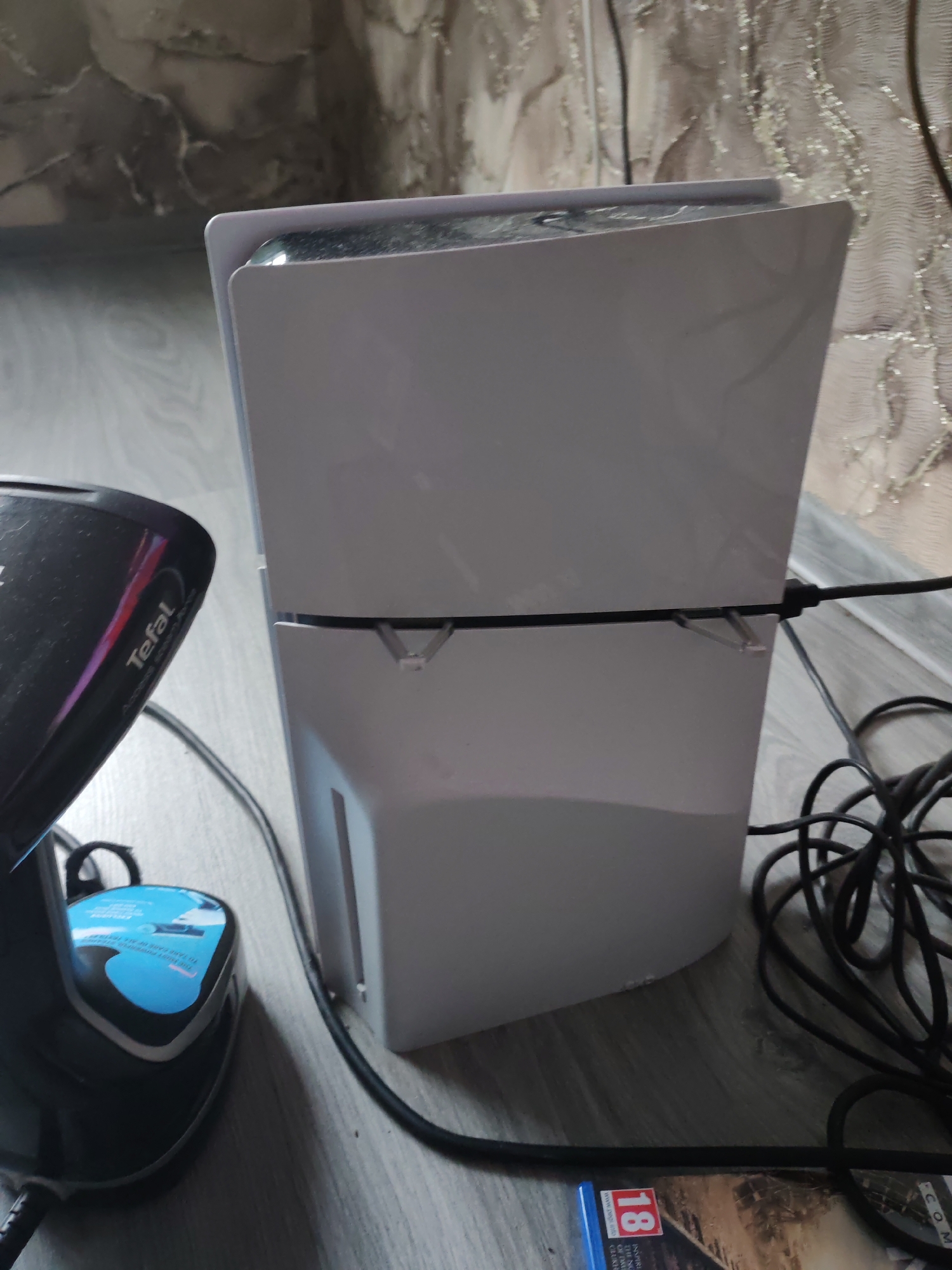 In Chulman, Russia, a white PlayStation 5 console stands vertically on a wooden floor. To the left of the console, a black Tefal steam generator iron is visible, its water tank displaying a blue label with text. Black power cables are connected to the console and lie on the floor. A portion of a video game case with the rating "18" is also present on the floor near the bottom right corner. The background shows a wall with textured wallpaper.