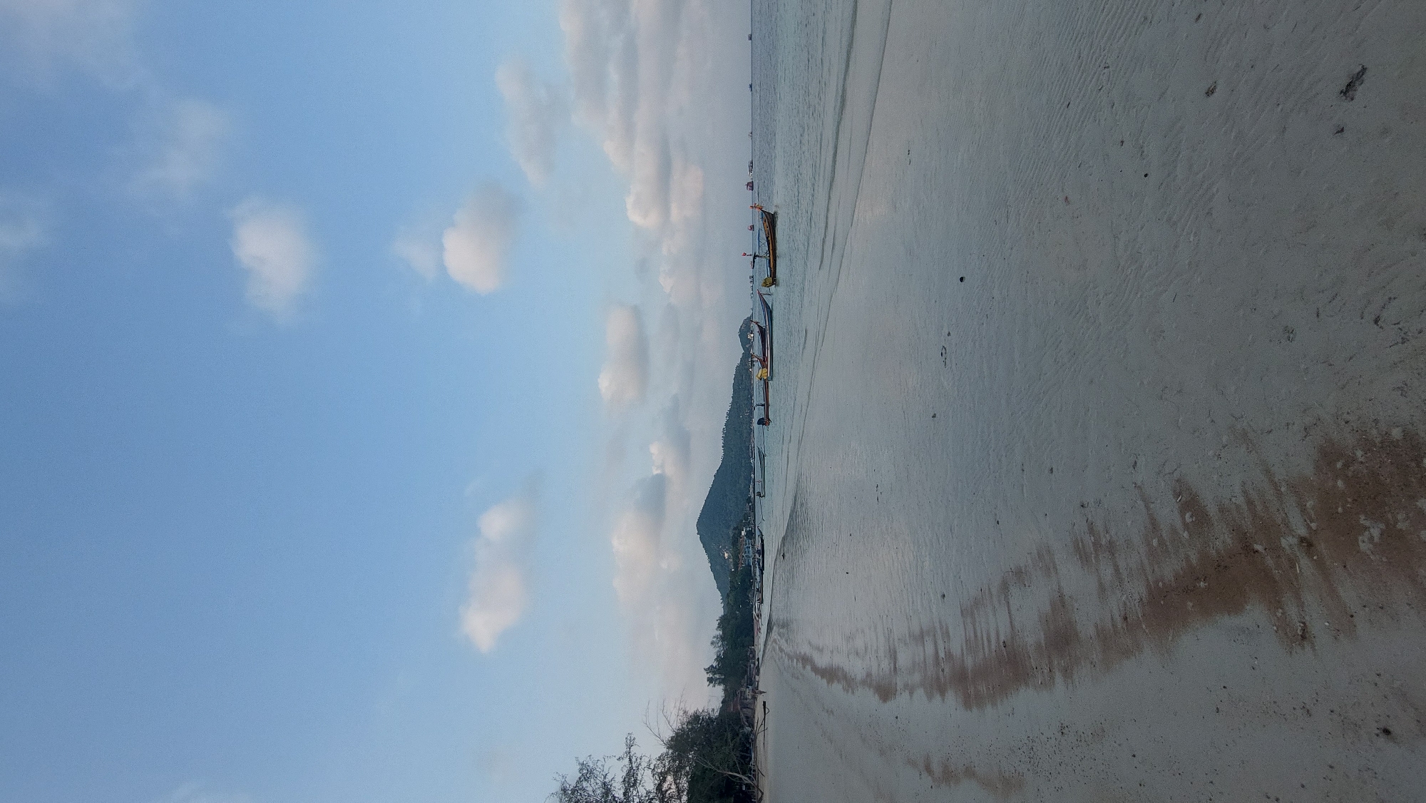 The image depicts a beach scene in Koh Tao, Thailand. The sand is light-colored and appears wet, suggesting a receding tide. There are several longtail boats anchored near the shoreline. In the background, a green mountain rises. The sky is blue with scattered white clouds. Vegetation is visible along the shoreline in the foreground.