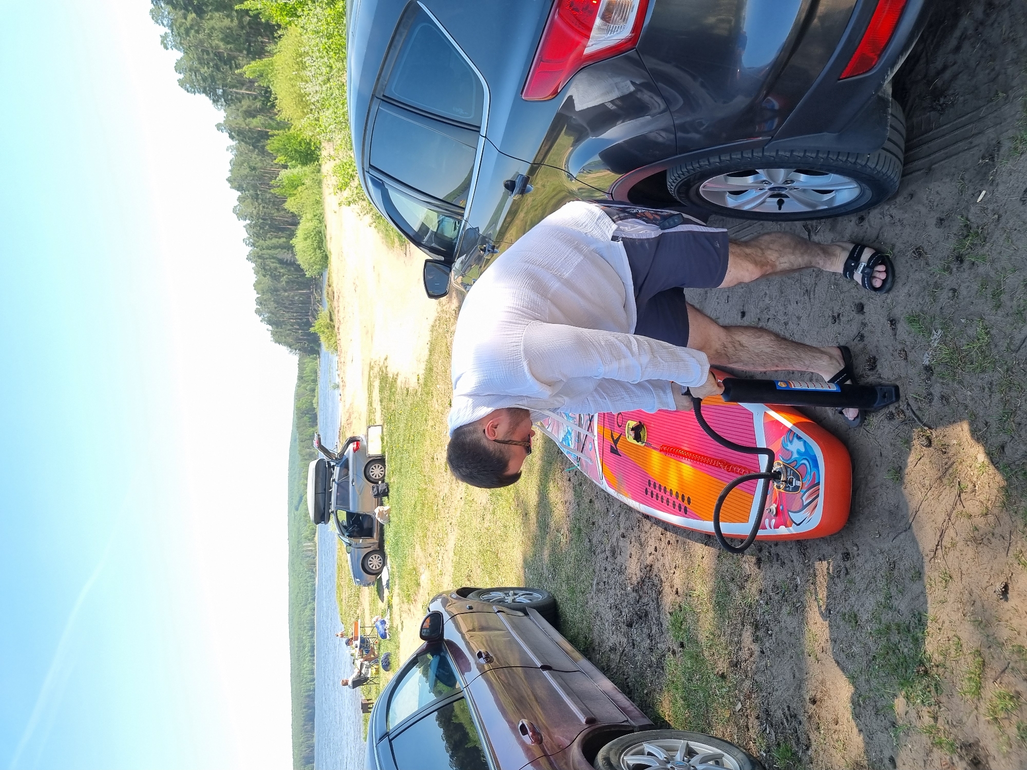 A man is inflating a red and orange paddleboard near two cars on a grassy shoreline. The location is near the city of Chaikovsky, Russia, adjacent to a body of water with other parked cars and people in the background. The man is wearing a white shirt, dark shorts, and sandals. One of the cars is dark gray and the other is maroon. The setting appears to be a recreational area, possibly a beach or lakeside park.
