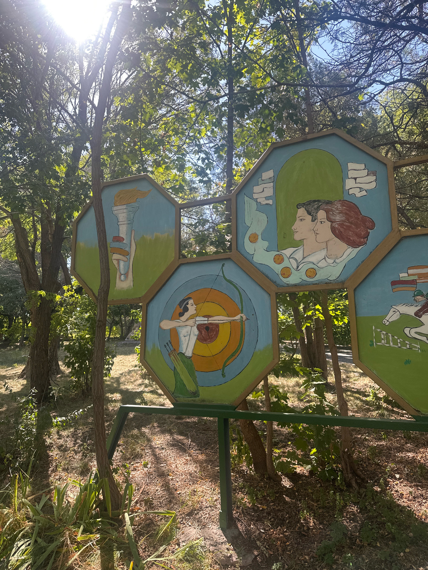 This is a medium shot, taken outdoors in a park-like setting with trees and dappled sunlight, showing a section of a colorful outdoor mural. The mural is composed of several hexagonal panels mounted on a green metal frame. The panels are painted with scenes related to sports and possibly the Olympics.
In the upper left panel, a hand holds a flaming torch against a light blue sky and green ground. To its right, in an upper panel, are the profiles of a man and a woman with their heads close together, surrounded by orange circles and light blue fabric. Below this, in a central panel, a woman is depicted in profile, aiming a bow and arrow at a target. In a panel to the right of the archer, a person on a white horse with a rider in a green helmet jumps over a hurdle. The style of the paintings is naive and illustrative. The overall scene suggests a public art installation likely located in a recreational area within the city of Gyumri, Armenia, possibly related to a cultural or sporting event. The ground is covered with dry leaves and grass, and young trees grow around the structure.