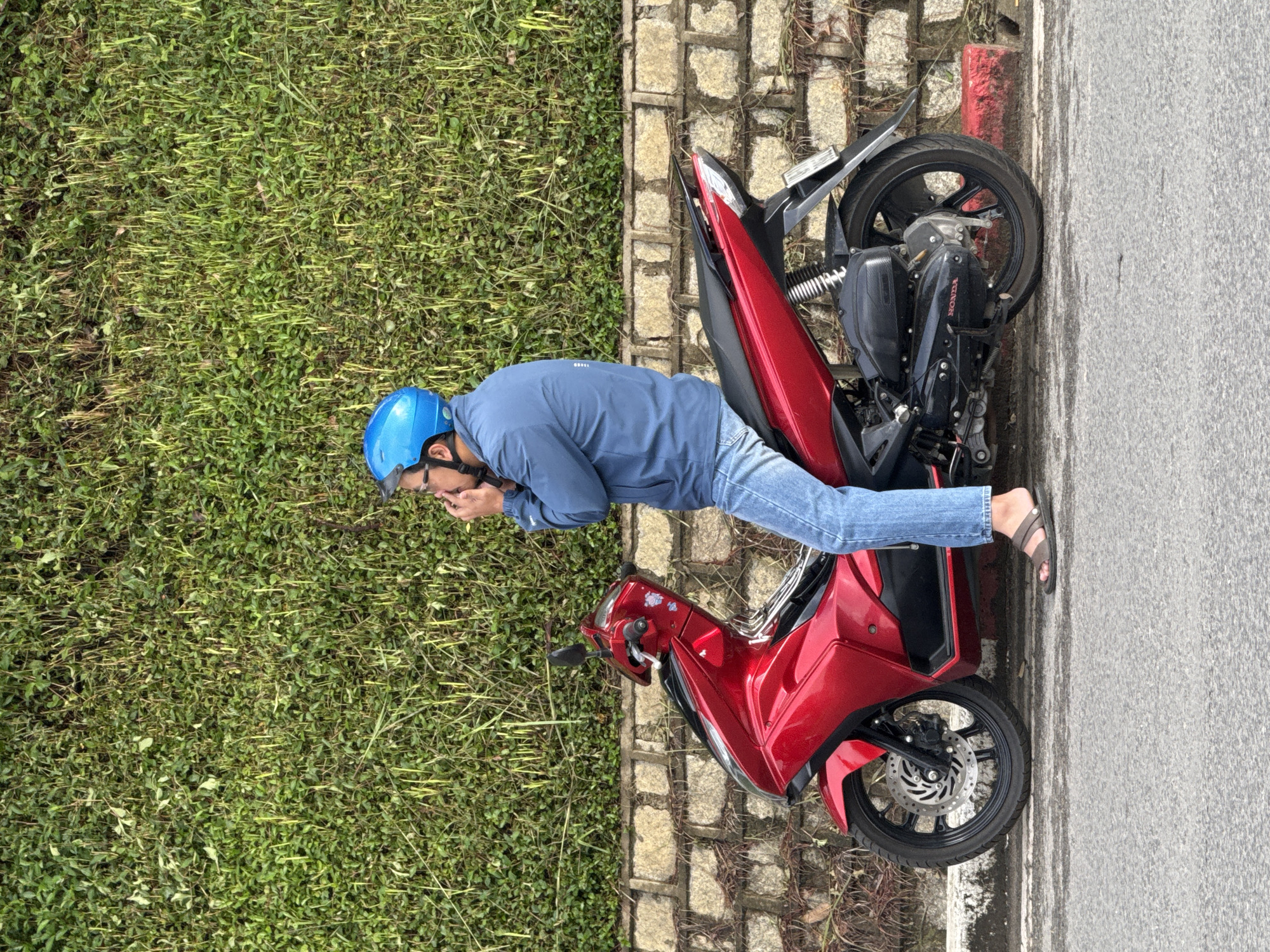 A side view of a man wearing a blue helmet, blue jacket, and blue jeans is seated on a red scooter. He is in the process of putting on or adjusting his helmet. The scooter is parked on the side of a road, next to a low stone wall covered in dense green foliage. The road surface is asphalt. Behind the foliage is a textured wall of stacked stones. A red curb is visible on the right side of the road. The setting appears to be an outdoor urban environment.